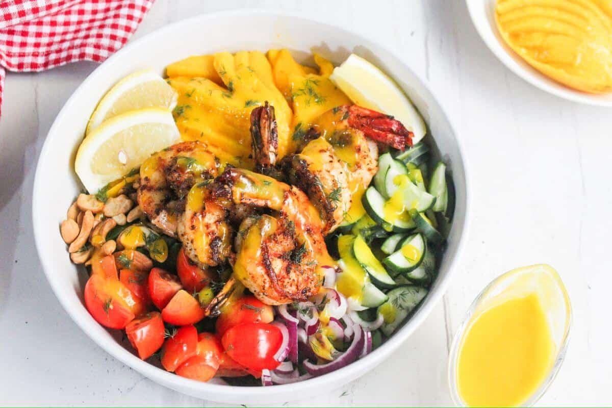 A bowl with grilled shrimp, sliced mango, lemon wedges, cherry tomatoes, cucumbers, red onions, peanuts, and greens, topped with a yellow sauce.