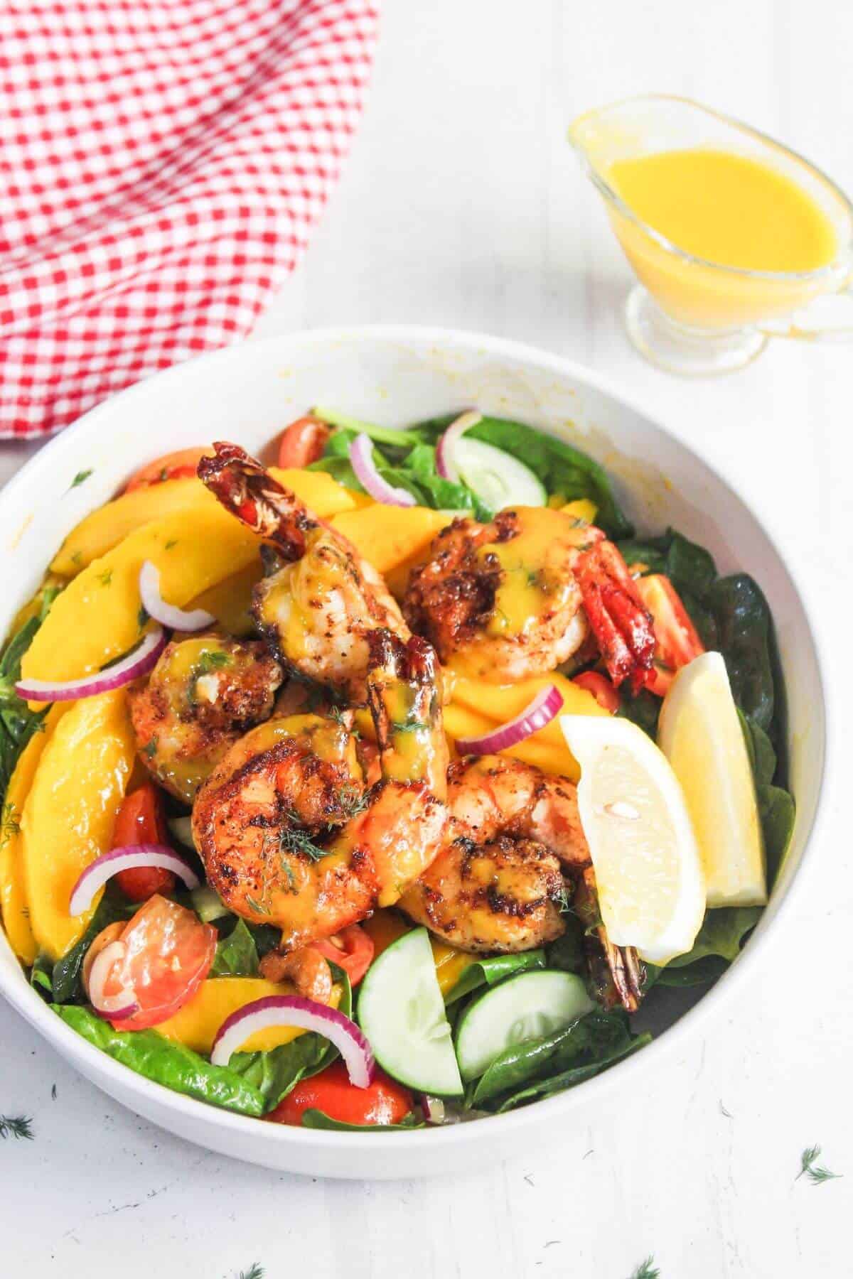 A bowl of salad with grilled shrimp, mango slices, spinach, cherry tomatoes, cucumber, red onion, lemon wedges, and a small jug of yellow dressing on the side.