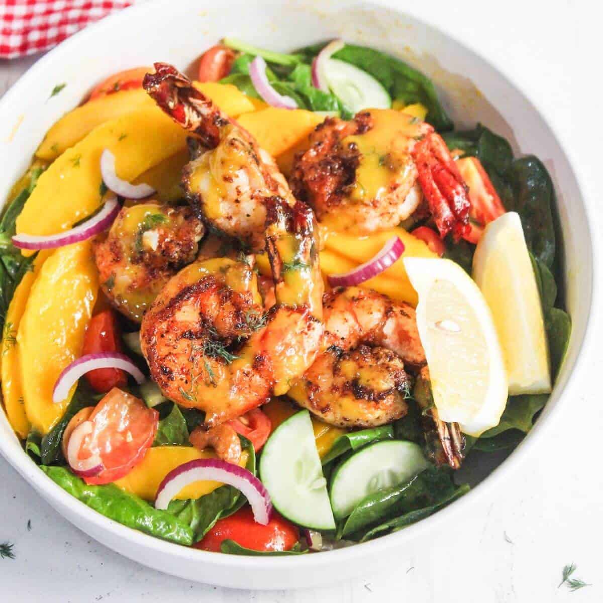 A bowl of grilled shrimp on a salad with mango slices, cherry tomatoes, cucumber, red onion, spinach, and lemon wedges.