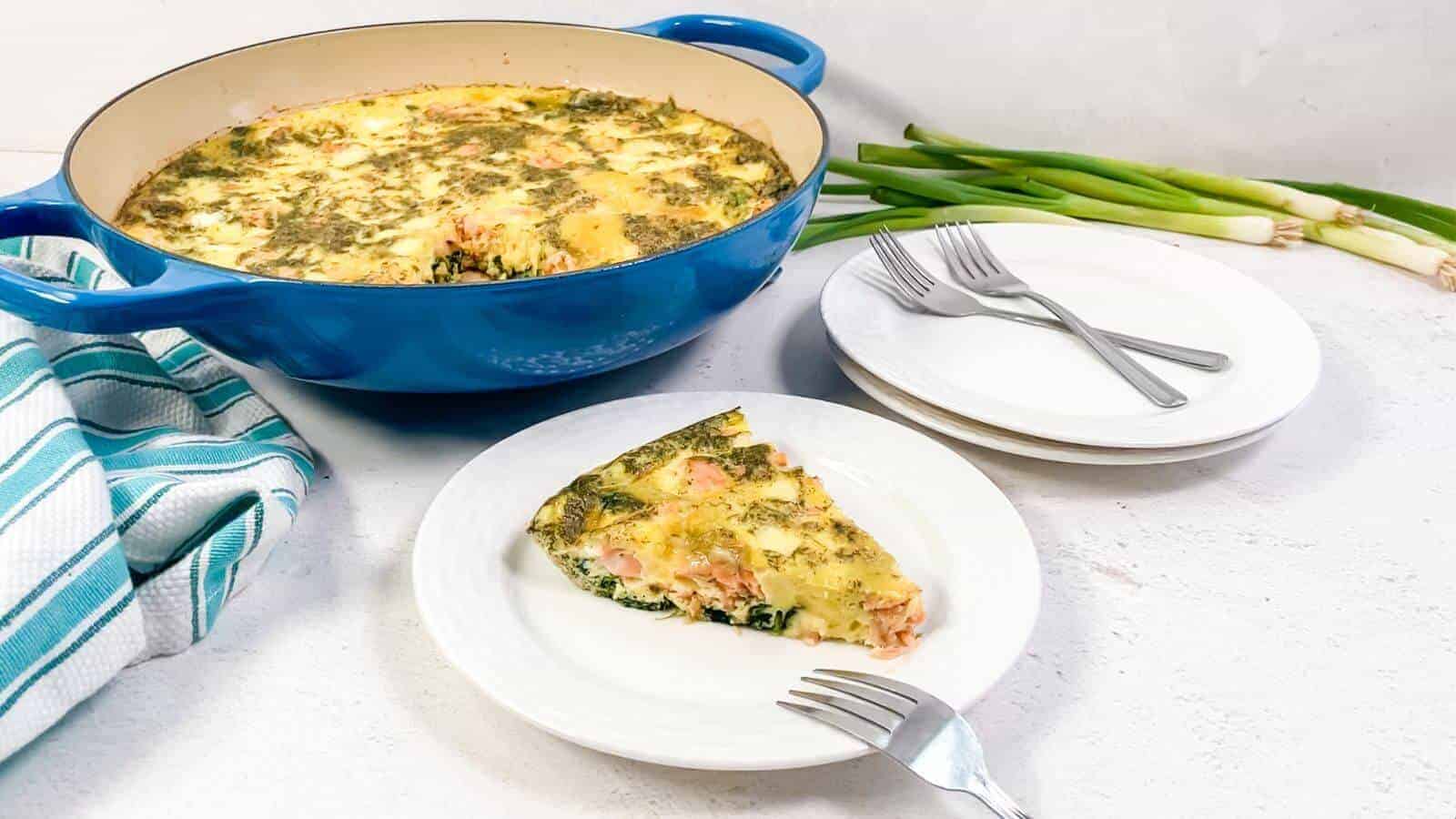 A slice of vegetable and cheese frittata is served on a white plate beside a blue baking dish, with stacked plates, forks, green onions, and a striped towel nearby.