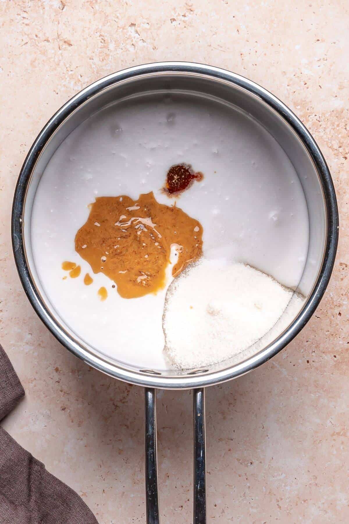 A saucepan containing coconut milk, peanut butter, vanilla extract, and granulated sugar on a beige countertop.