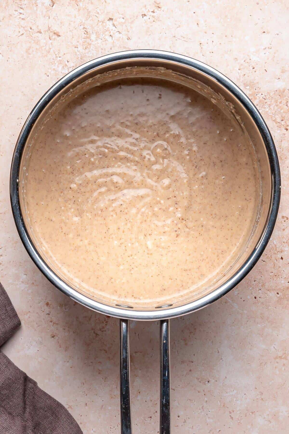A metal saucepan filled with a light brown, creamy sauce or batter sitting on a beige surface.