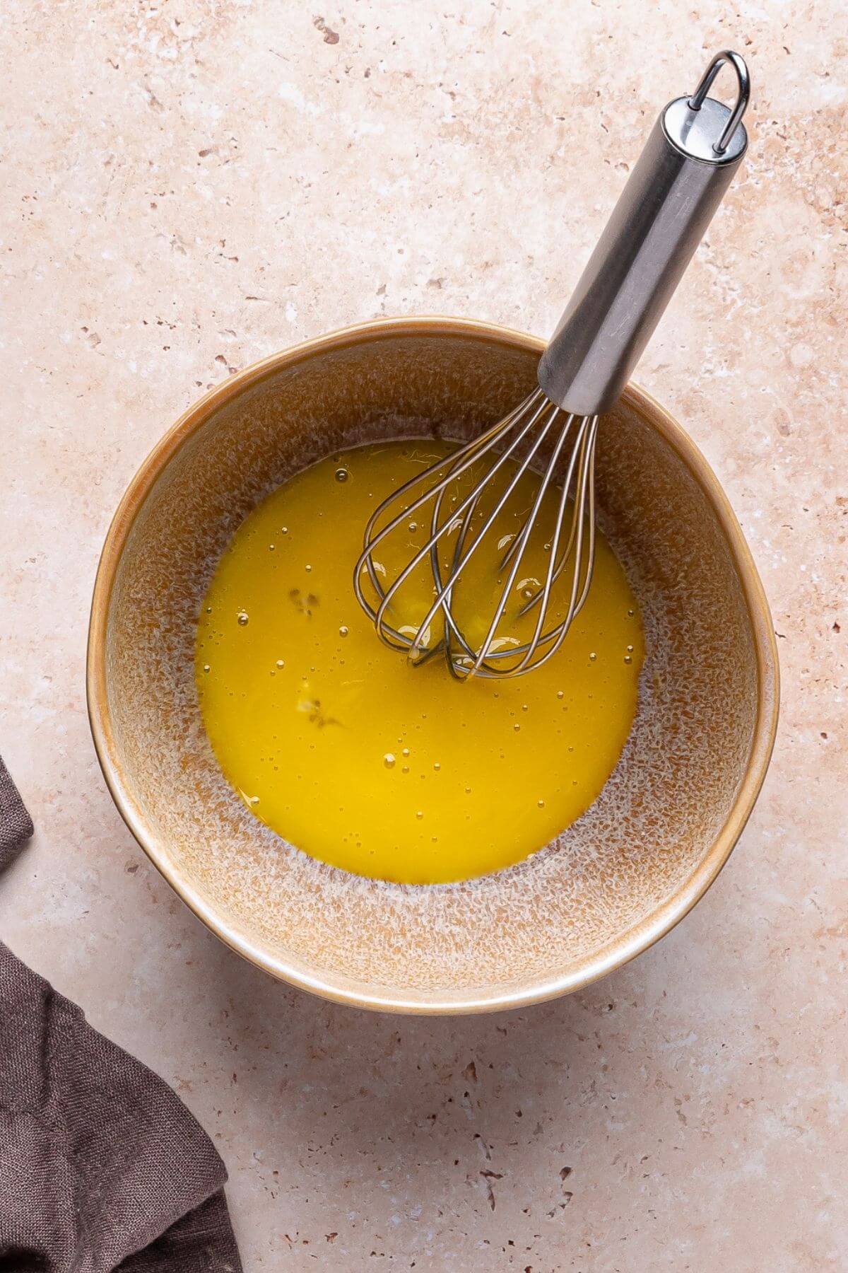 A whisk rests in a ceramic bowl containing beaten eggs on a light countertop, with a brown cloth nearby.