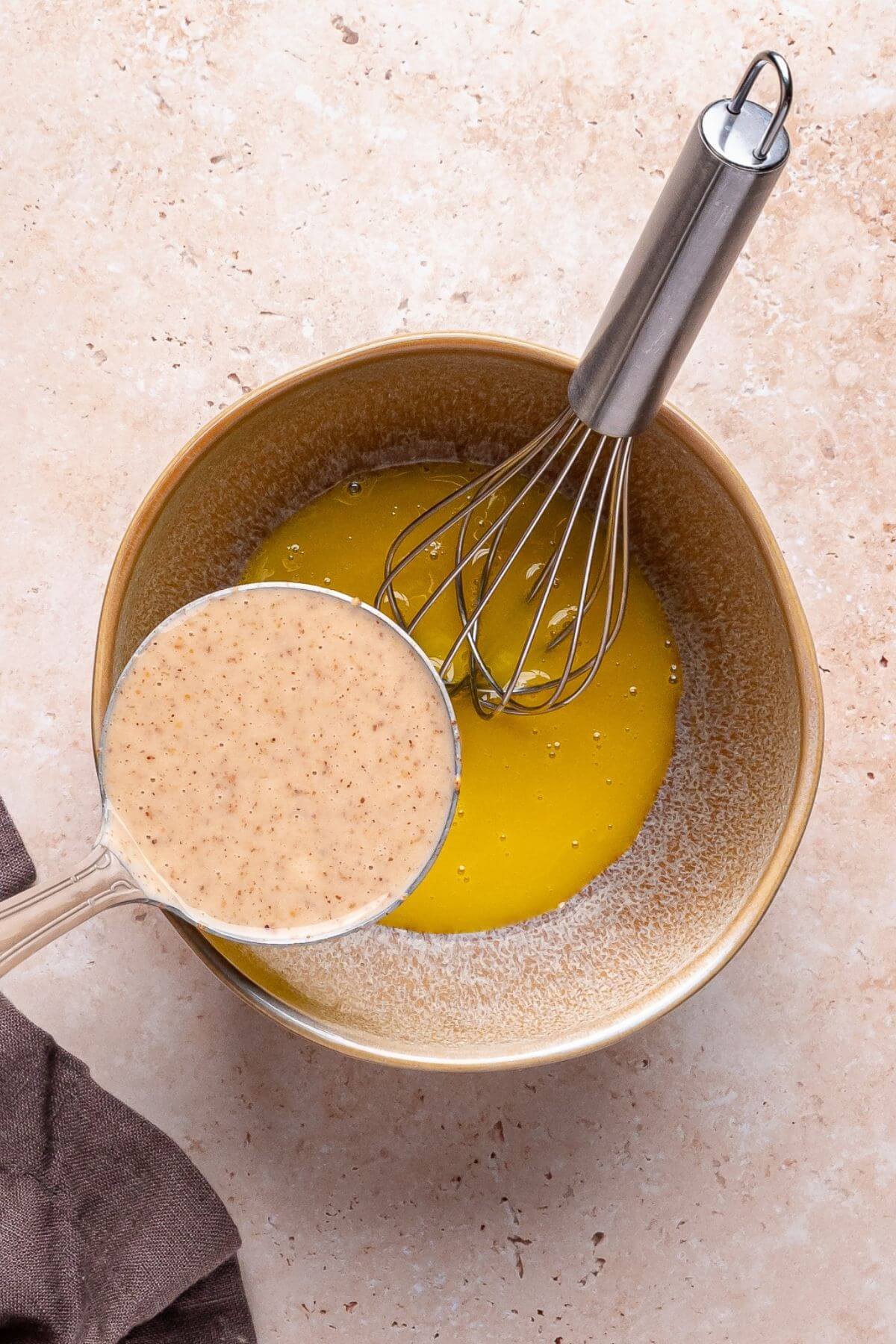 A whisk rests in a bowl containing melted butter, while a measuring cup filled with a creamy mixture is held above the bowl on a light countertop.