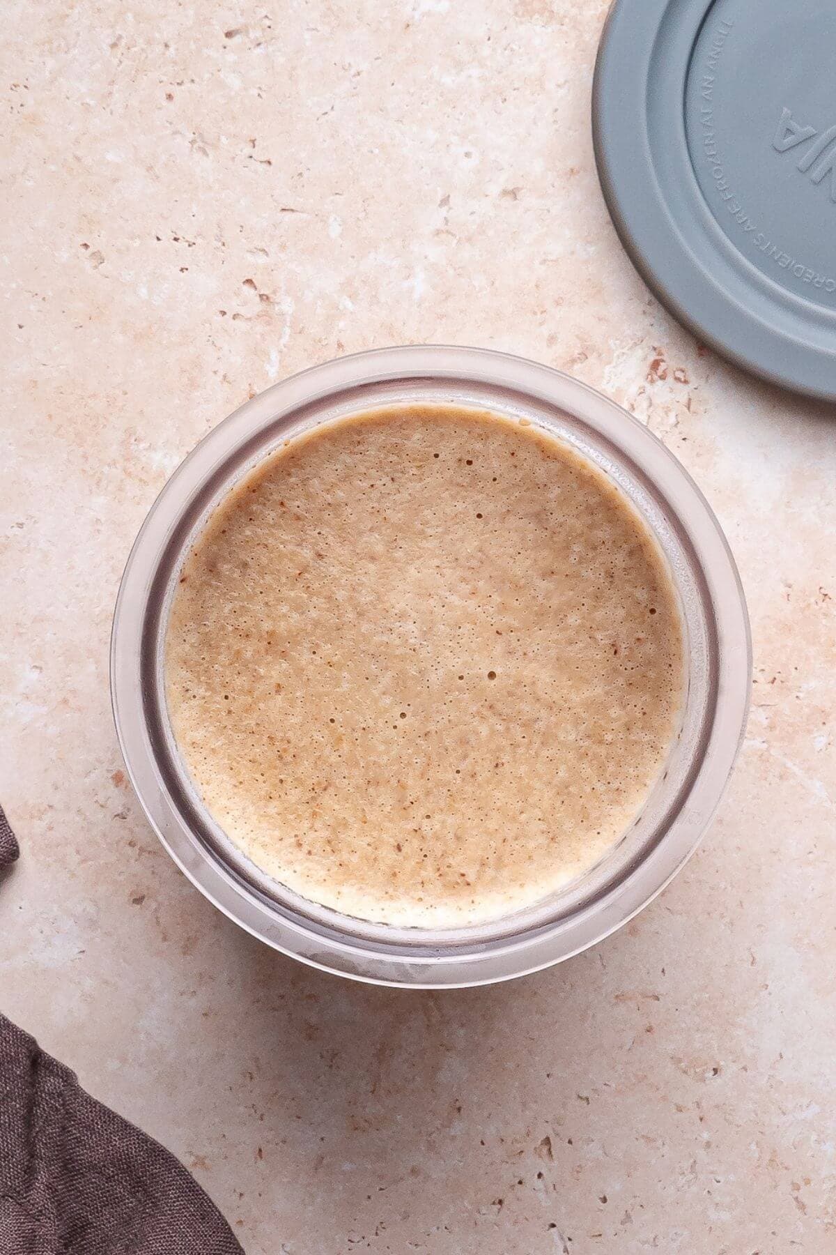 A glass container filled with frothy brown liquid sits on a beige surface next to a gray lid and a brown cloth.