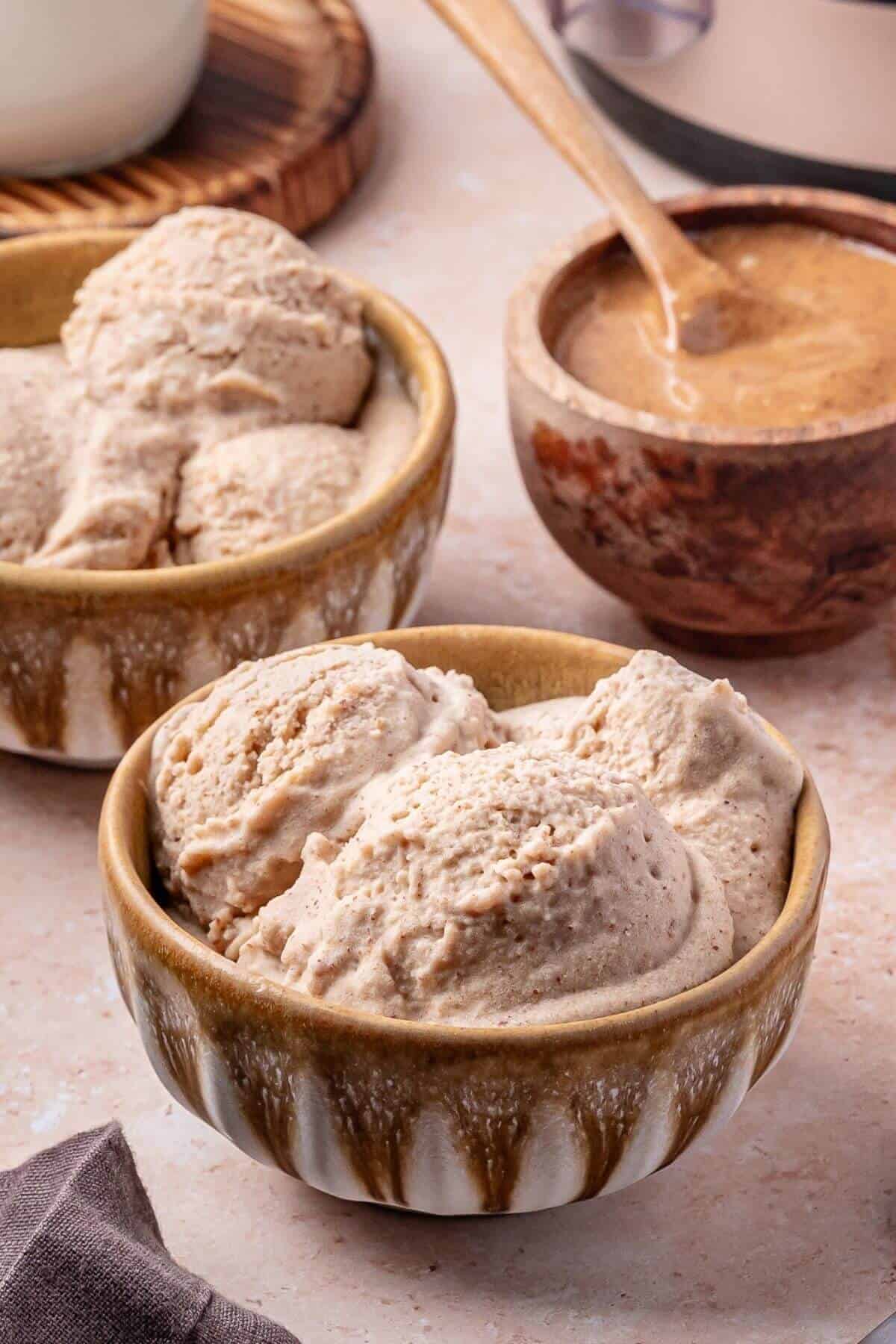 A ceramic bowl filled with three scoops of light brown ice cream sits on a countertop, with another bowl of ice cream and a small bowl of sauce in the background.