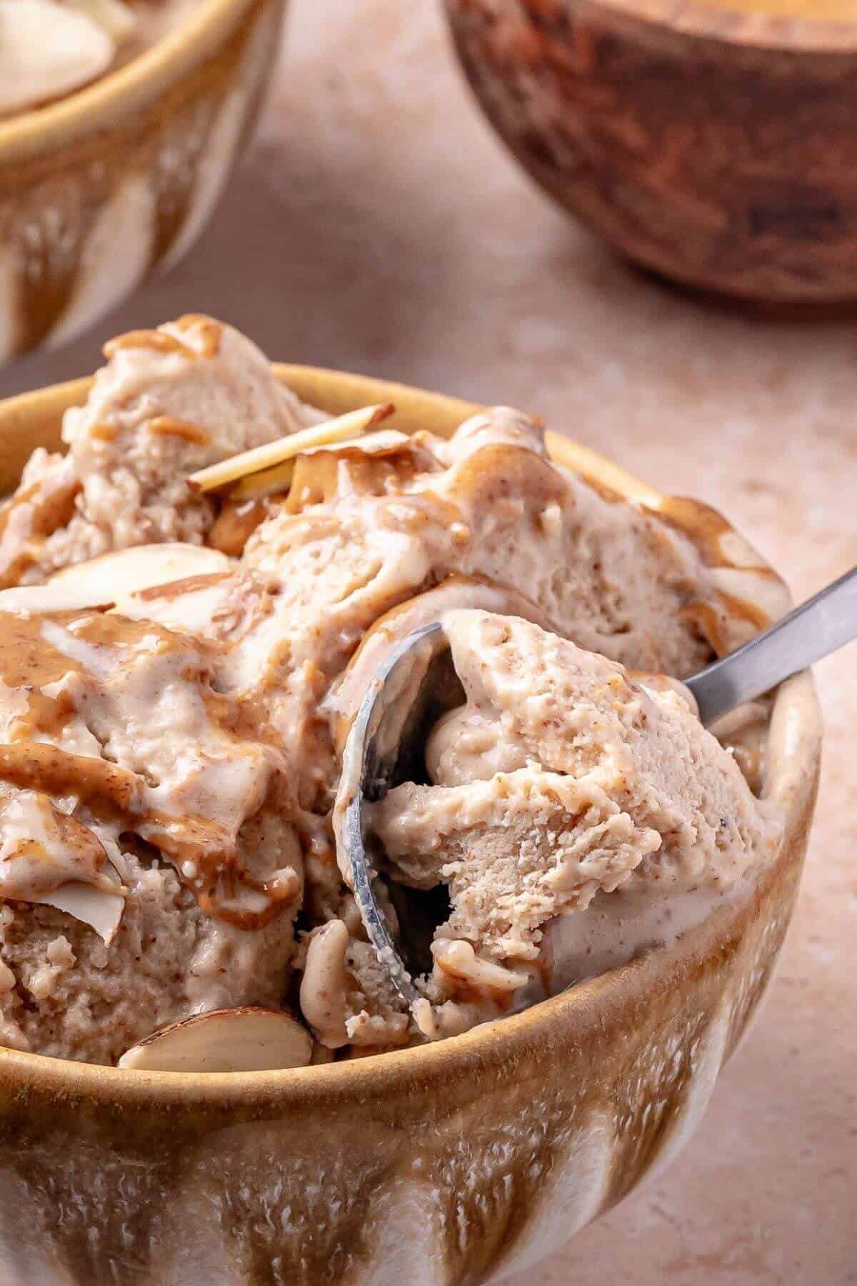 A close-up of a bowl filled with creamy ice cream, topped with sliced almonds and caramel sauce, with a spoon scooping some out.