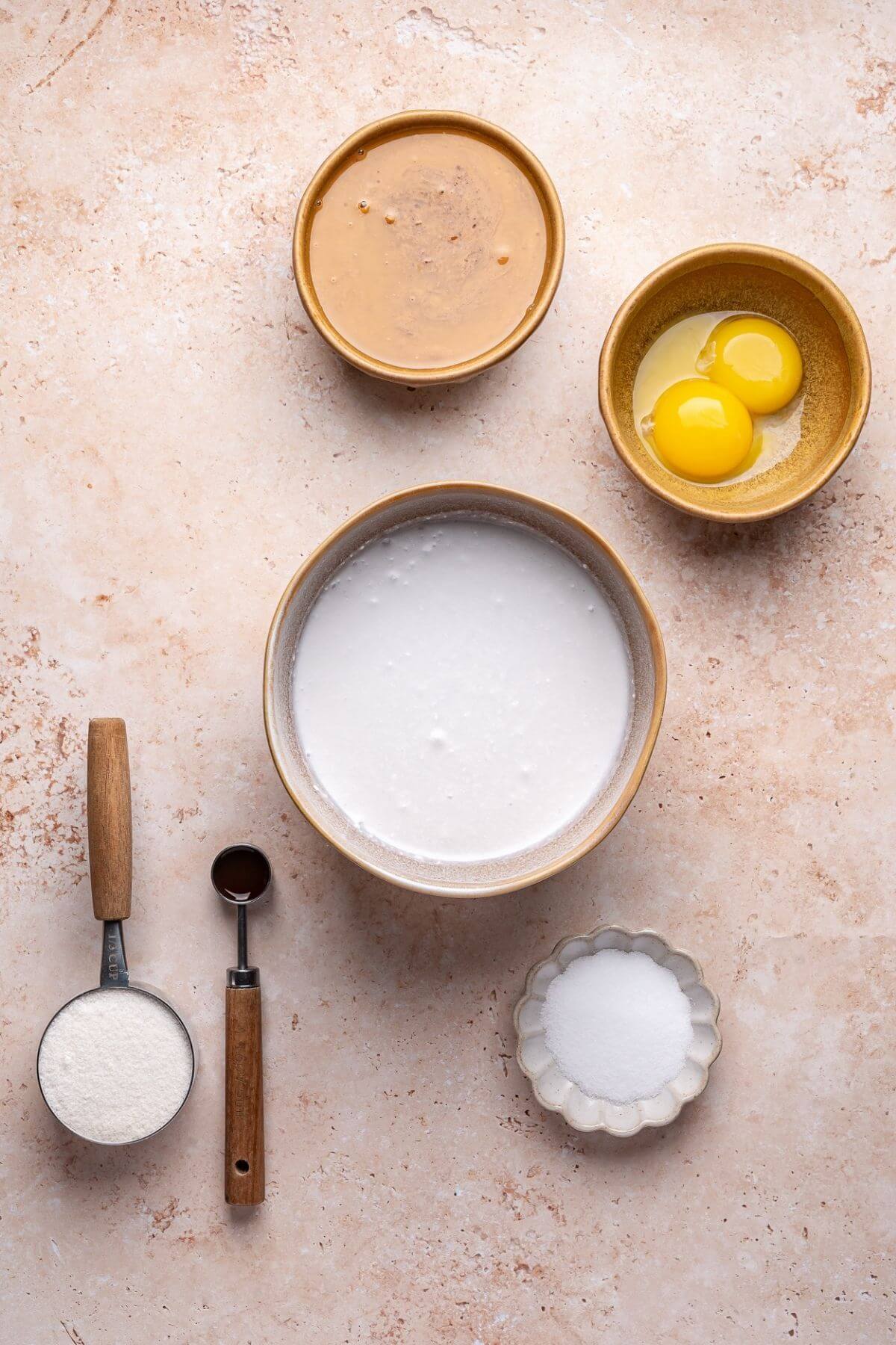 Five bowls and utensils on a light surface containing peanut butter, two eggs, coconut milk, flour, vanilla extract, and sugar as separate baking ingredients.