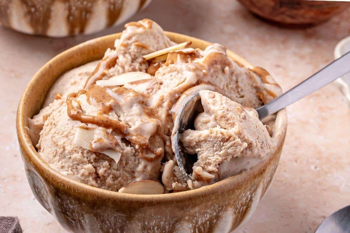 A bowl of ice cream topped with sliced almonds and a drizzle of brown sauce, with a spoon in the bowl.