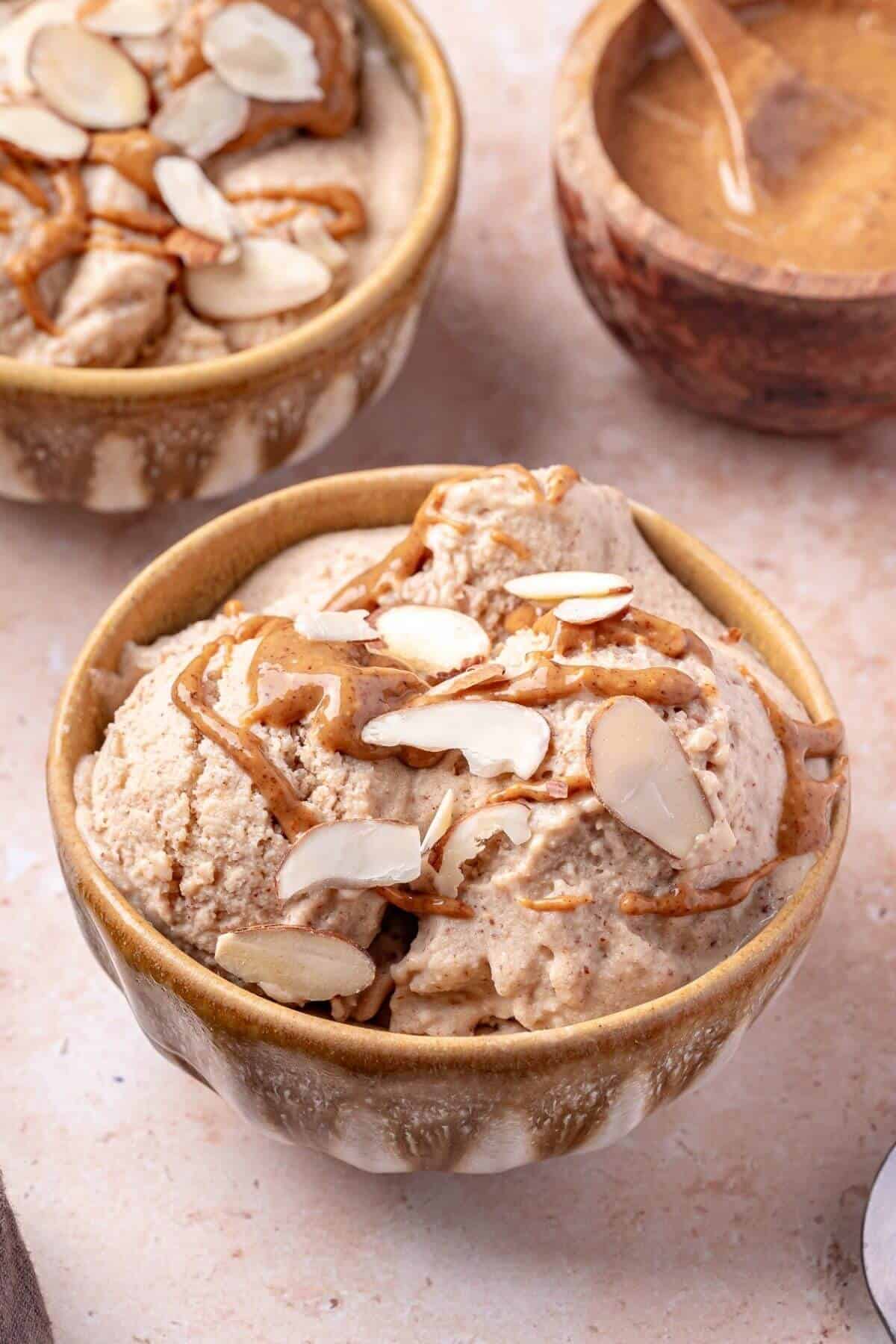 A bowl of ice cream topped with almond slices and a drizzle of caramel sauce, with a wooden bowl of sauce and another bowl of ice cream in the background.