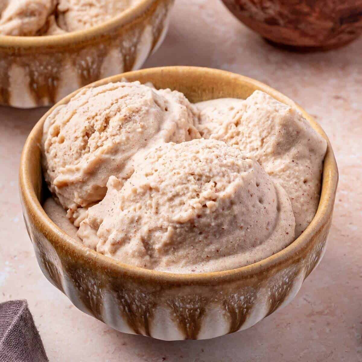 A ceramic bowl filled with three scoops of light brown ice cream sits on a light-colored surface.