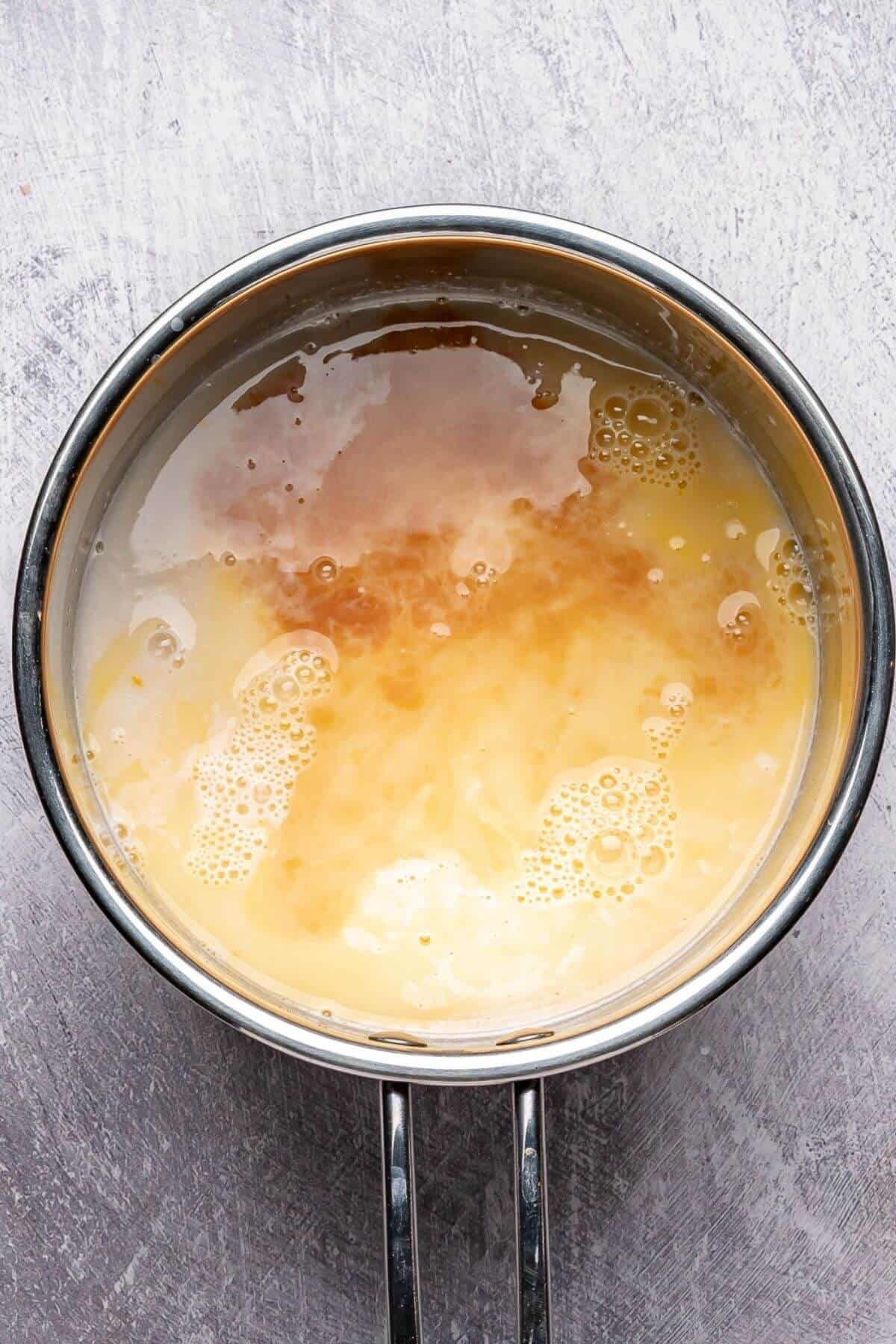 A saucepan containing an even mixture of liquids, with a light tan color and some bubbles forming on the surface.
