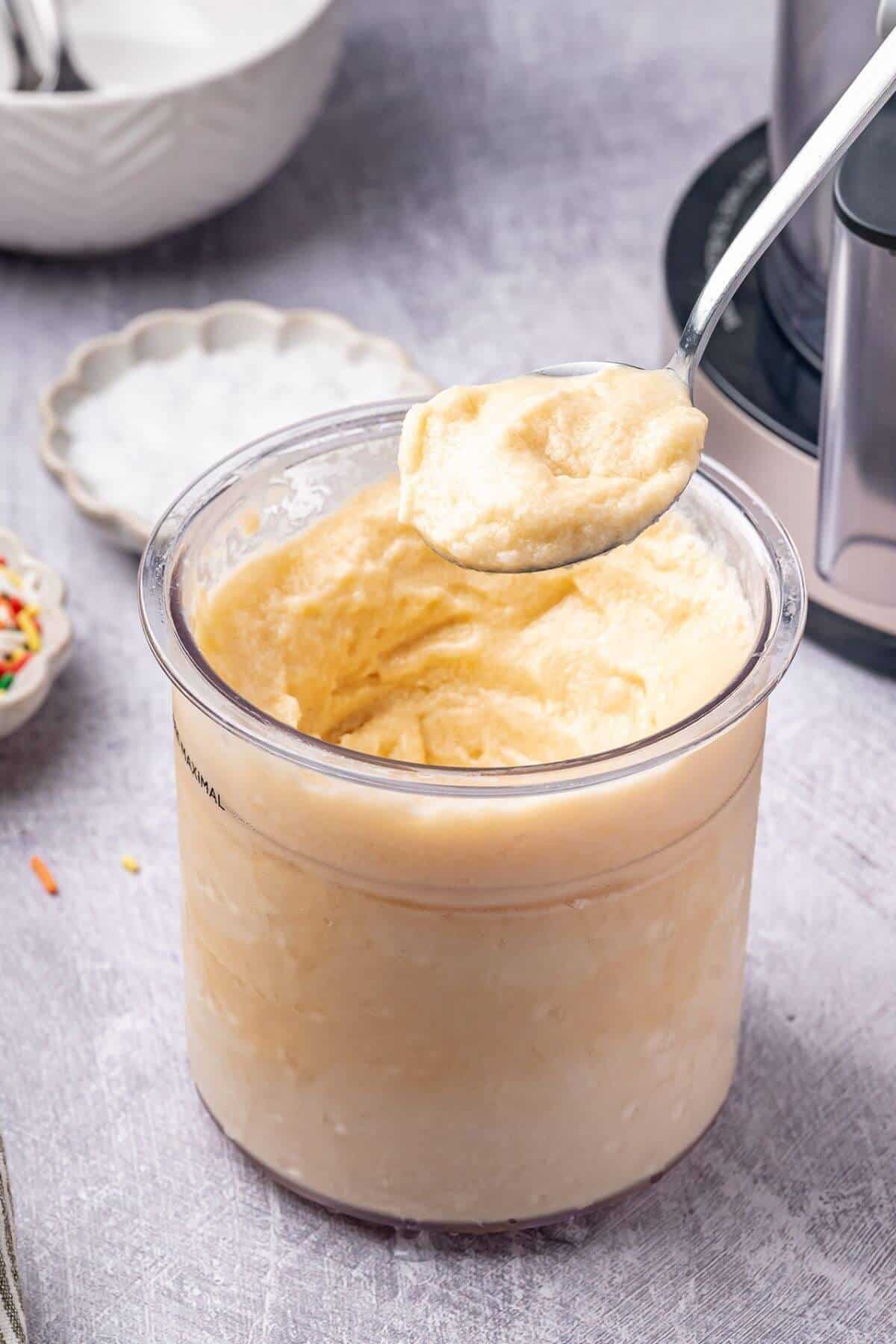 A spoon holding vanilla soft serve ice cream above a transparent container filled with more of the same ice cream, placed on a light gray surface.