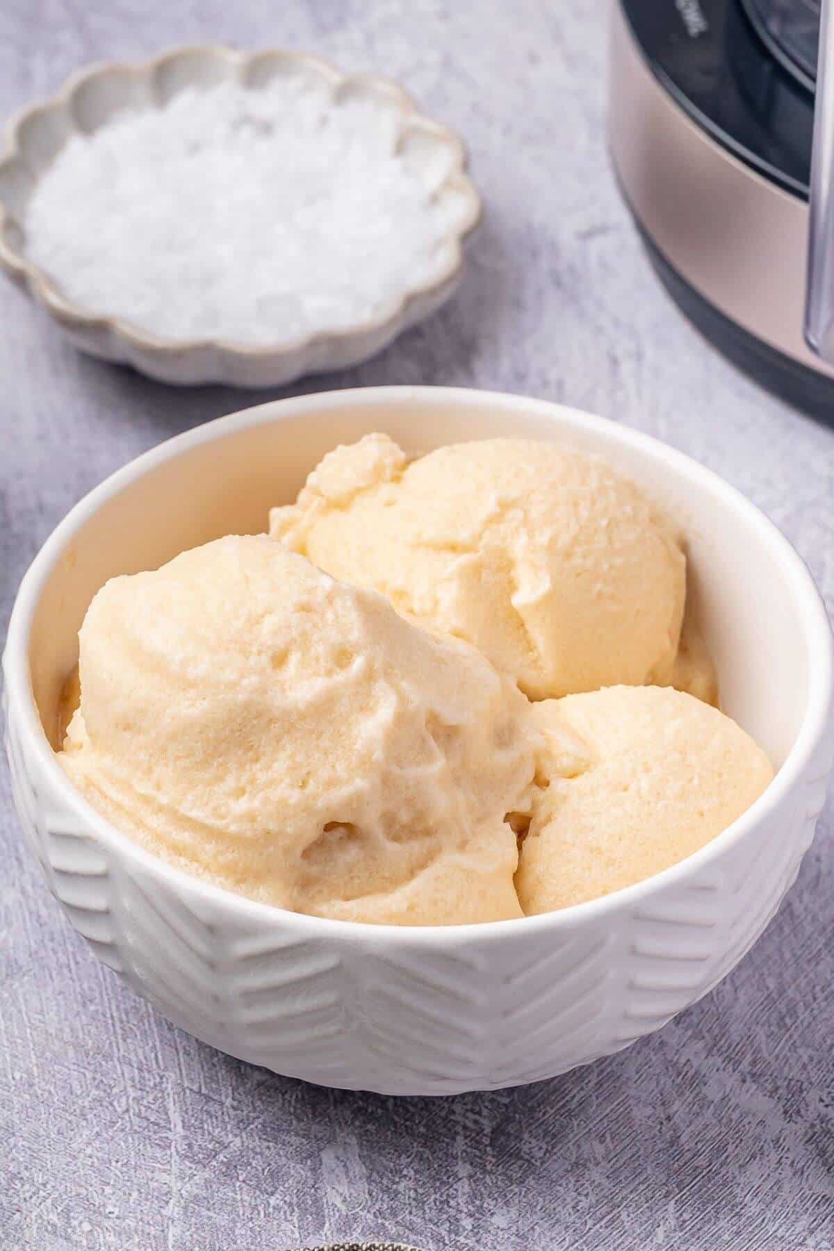 A white bowl filled with three scoops of vanilla ice cream sits on a light gray surface, with a dish of salt and a kitchen appliance nearby.