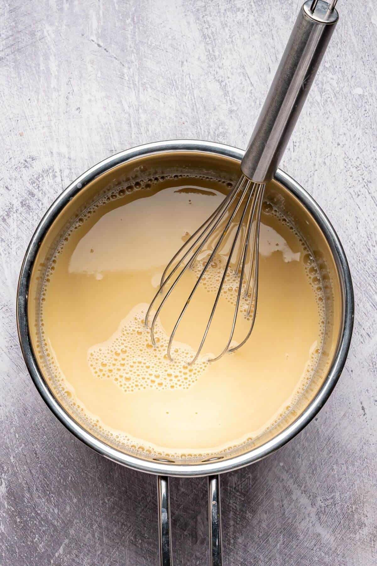 A metal whisk resting in a saucepan filled with a light brown, creamy liquid on a textured gray surface.