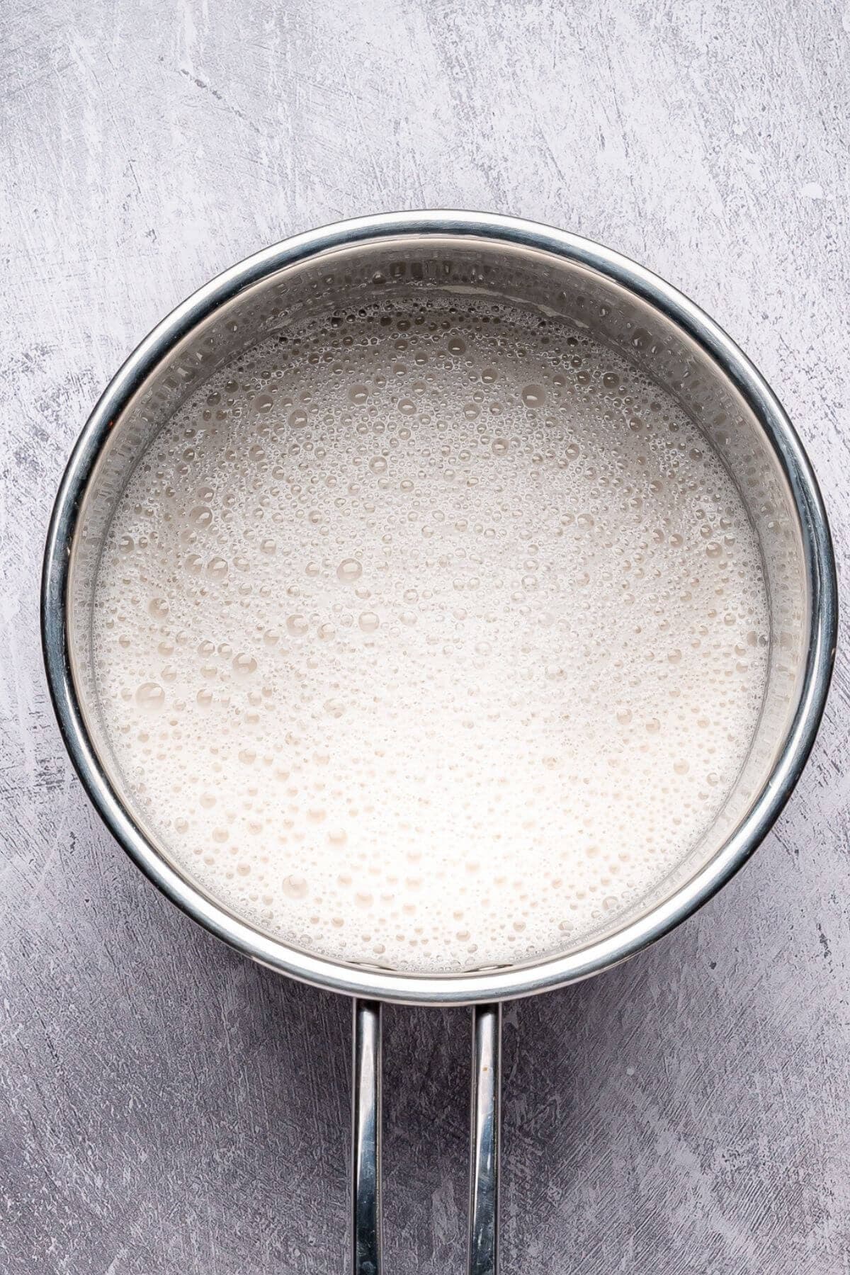 A stainless steel saucepan filled with a frothy, off-white liquid sits on a textured gray surface.
