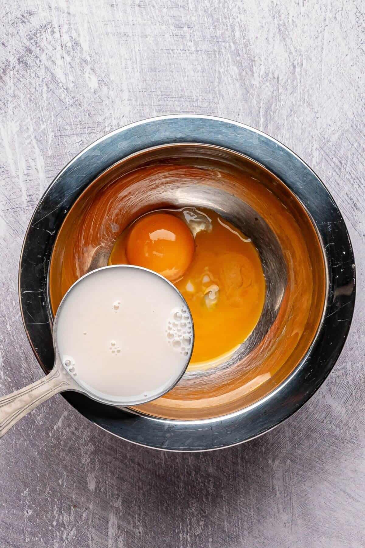A metal bowl with two cracked eggs inside, next to a metal measuring cup filled with milk, on a light-colored surface.