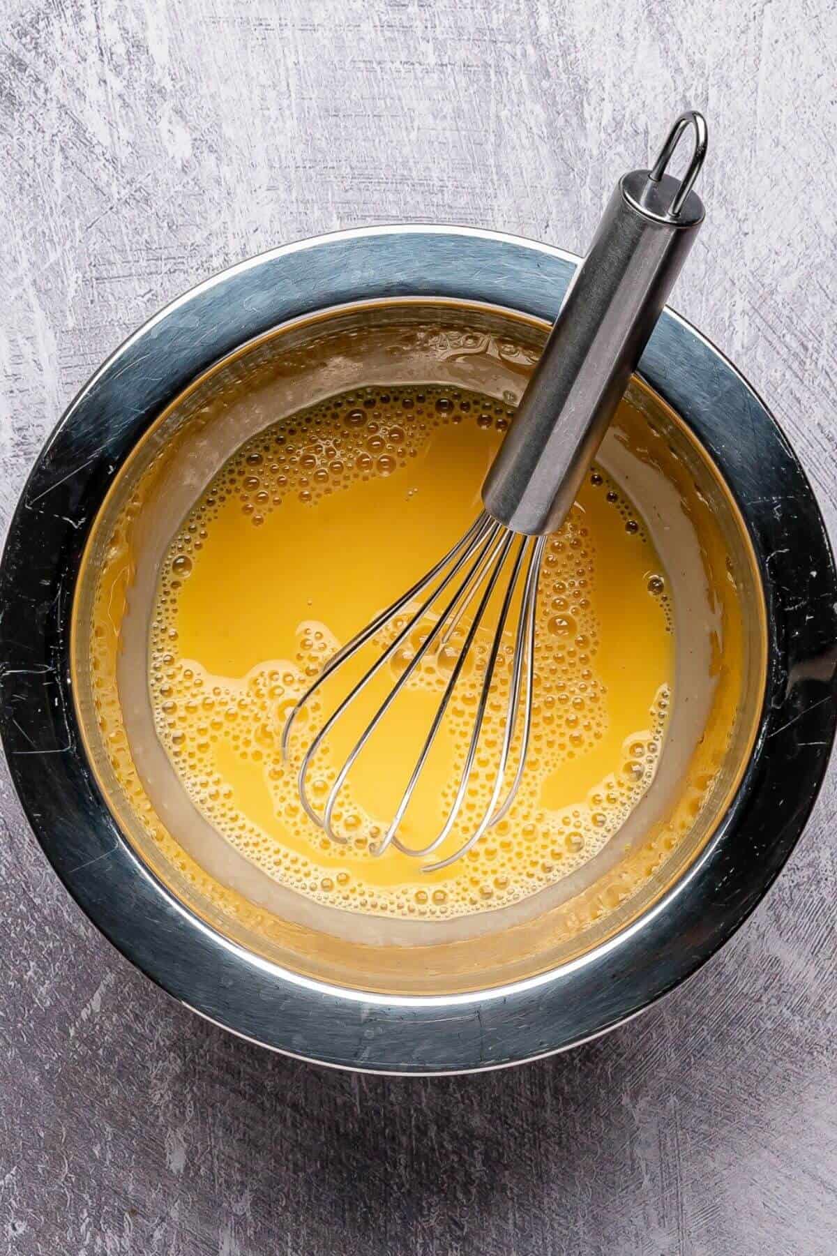 A metal whisk rests in a stainless steel bowl filled with a yellow liquid mixture on a gray textured surface.