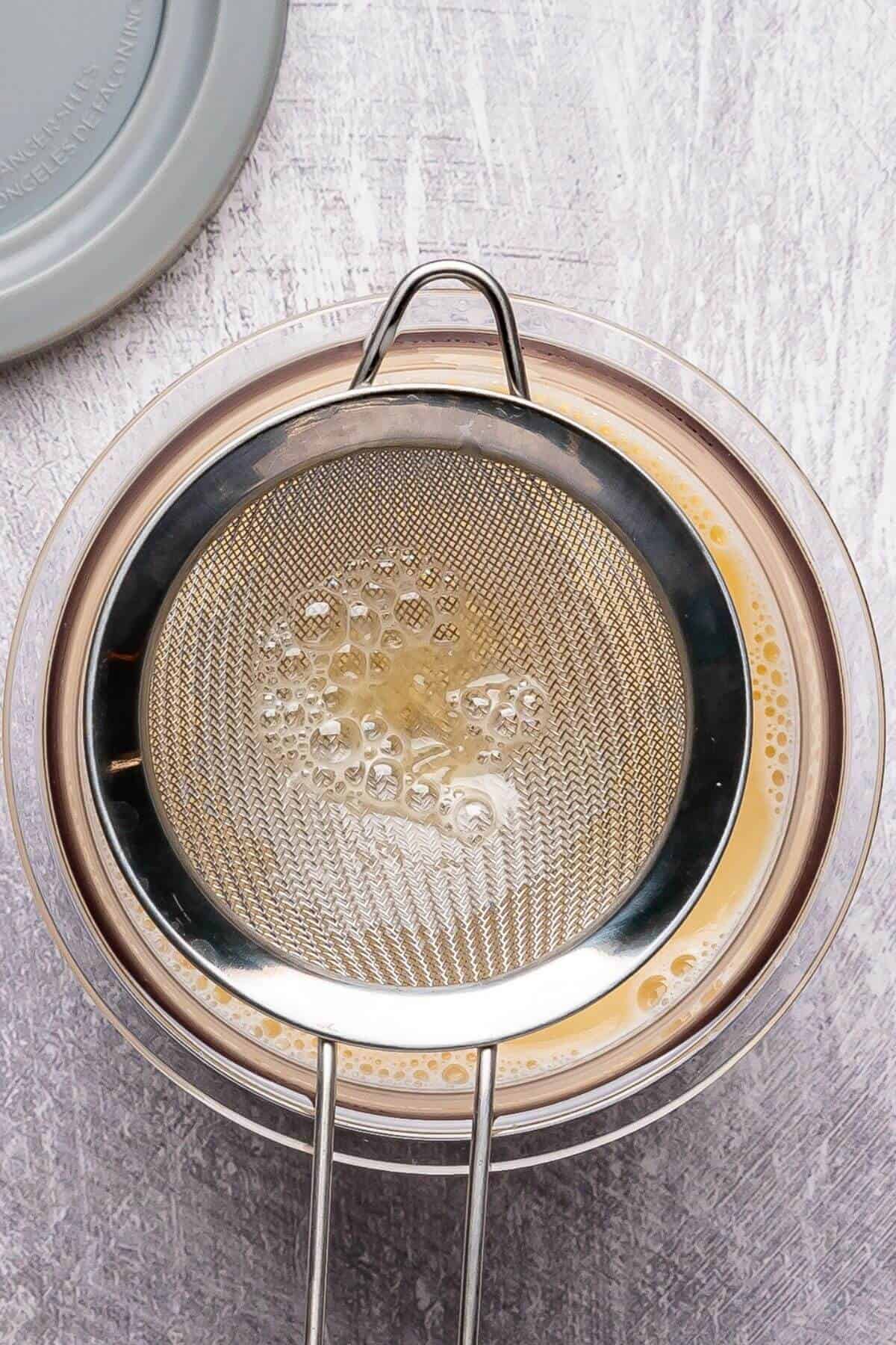 A metal strainer sits over a clear bowl, filtering a yellowish liquid on a light gray textured surface.