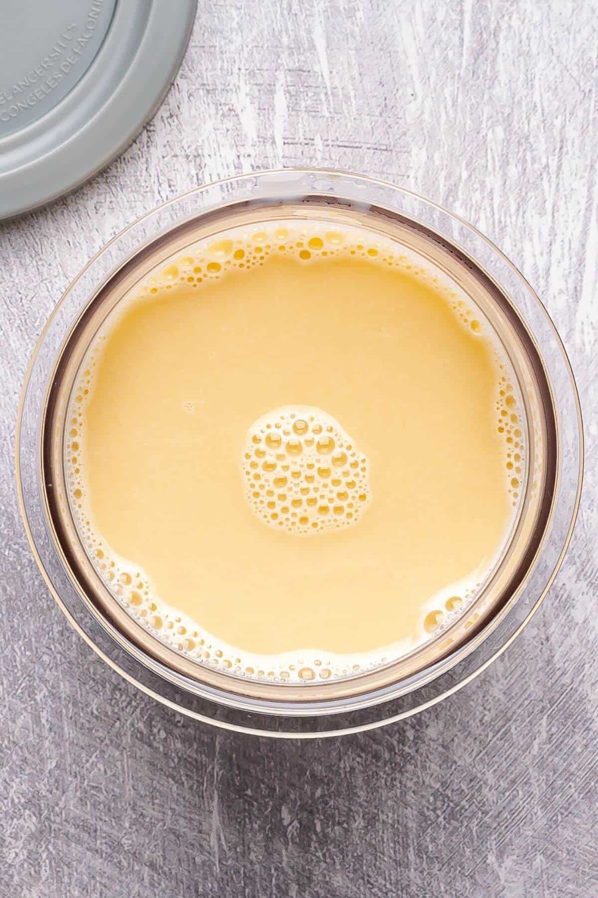 A clear bowl filled with creamy yellow liquid, possibly a sauce or blended mixture, sits on a gray textured surface with a plastic lid partially visible.