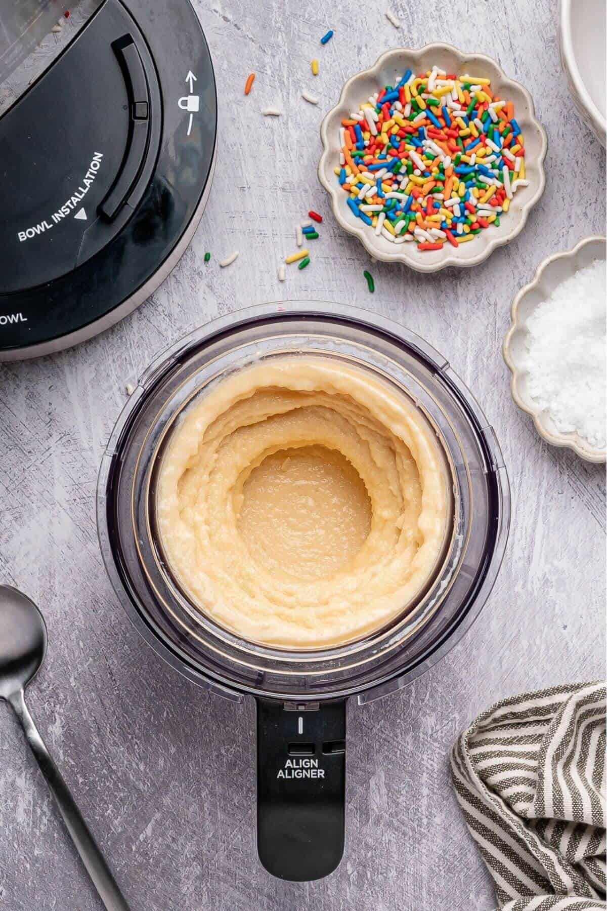 A food processor with blended batter inside sits on a countertop next to a bowl of rainbow sprinkles, a bowl of white powder, a spoon, and a striped towel.