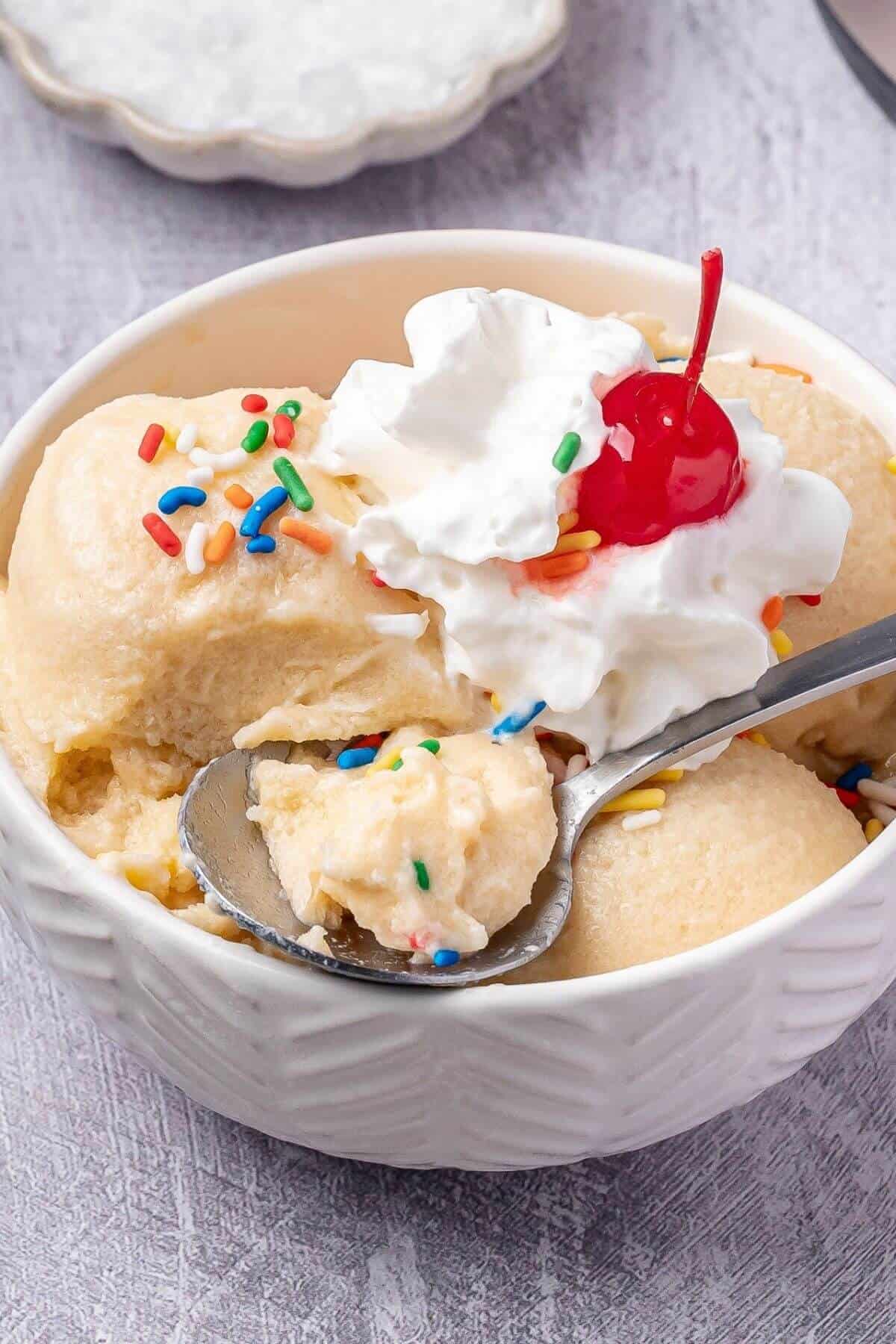 A bowl of ice cream topped with whipped cream, a cherry, and colorful sprinkles; a spoon rests in the bowl with a scoop of ice cream.