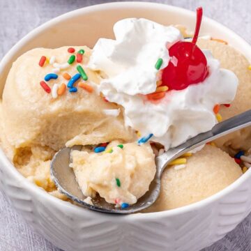 A bowl of vanilla ice cream topped with whipped cream, a cherry, and colorful sprinkles, with a spoonful being lifted.