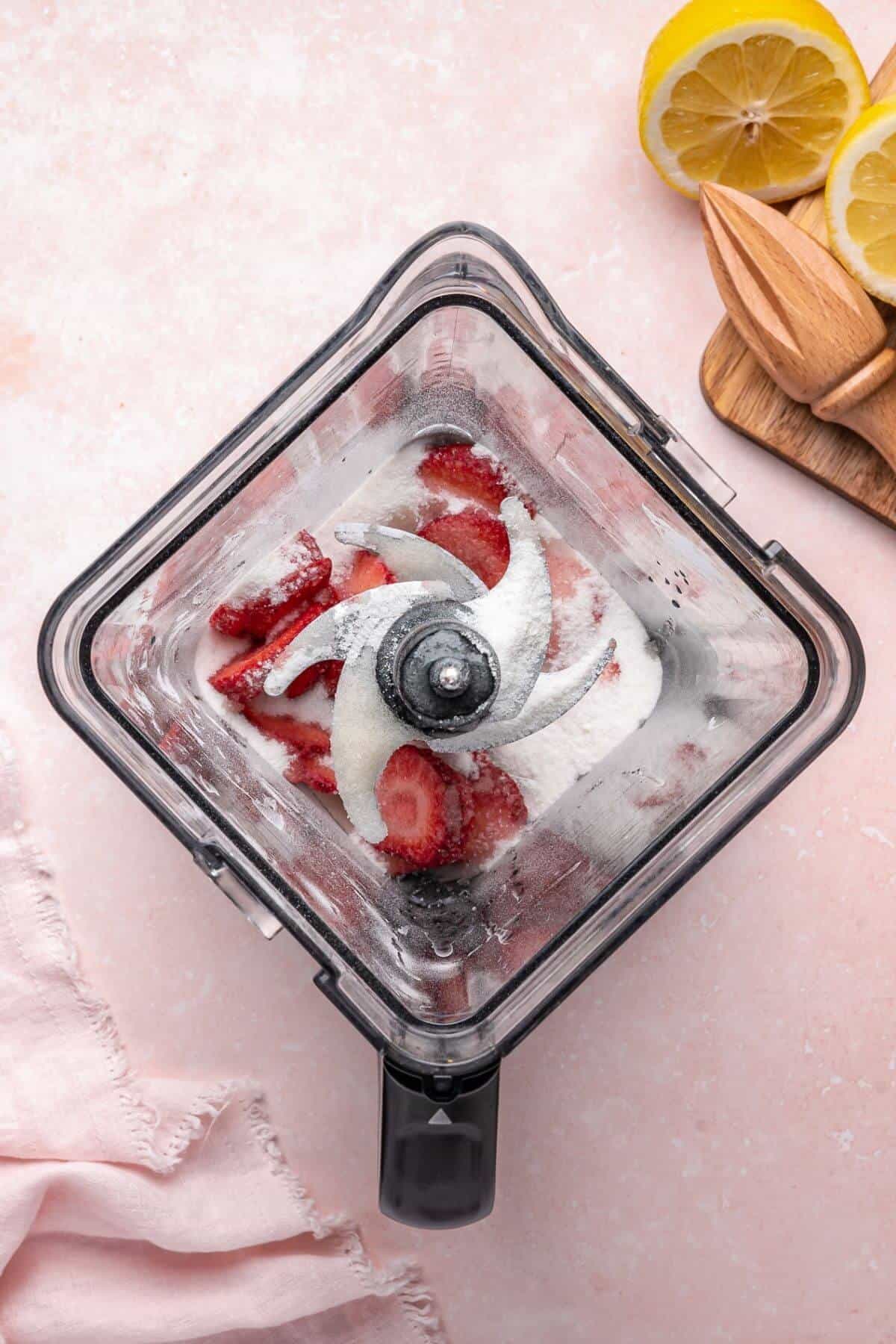 Overhead view of a blender containing sliced strawberries and sugar, with halved lemons and a wooden citrus juicer nearby on a pink surface.
