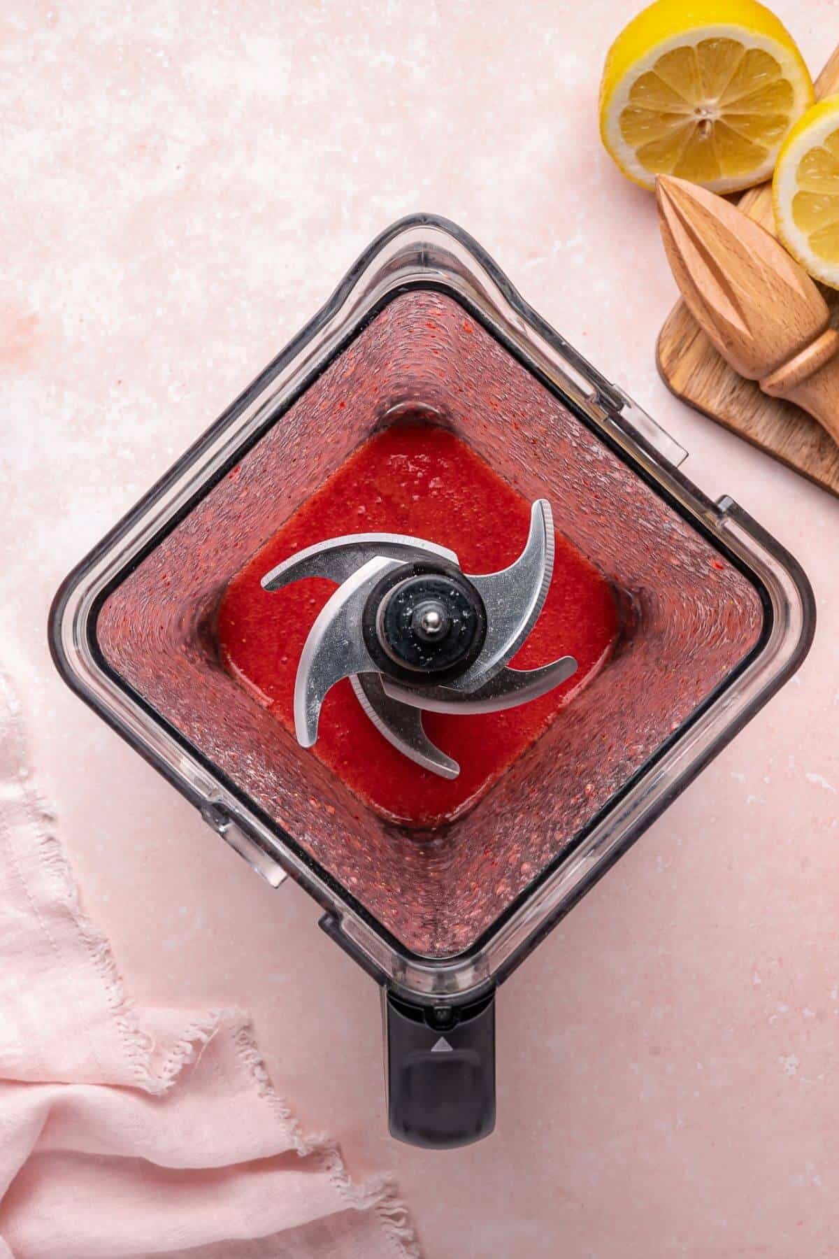A blender jar filled with a red liquid mixture, viewed from above, with sliced lemons and a wooden reamer nearby on a light pink surface.
