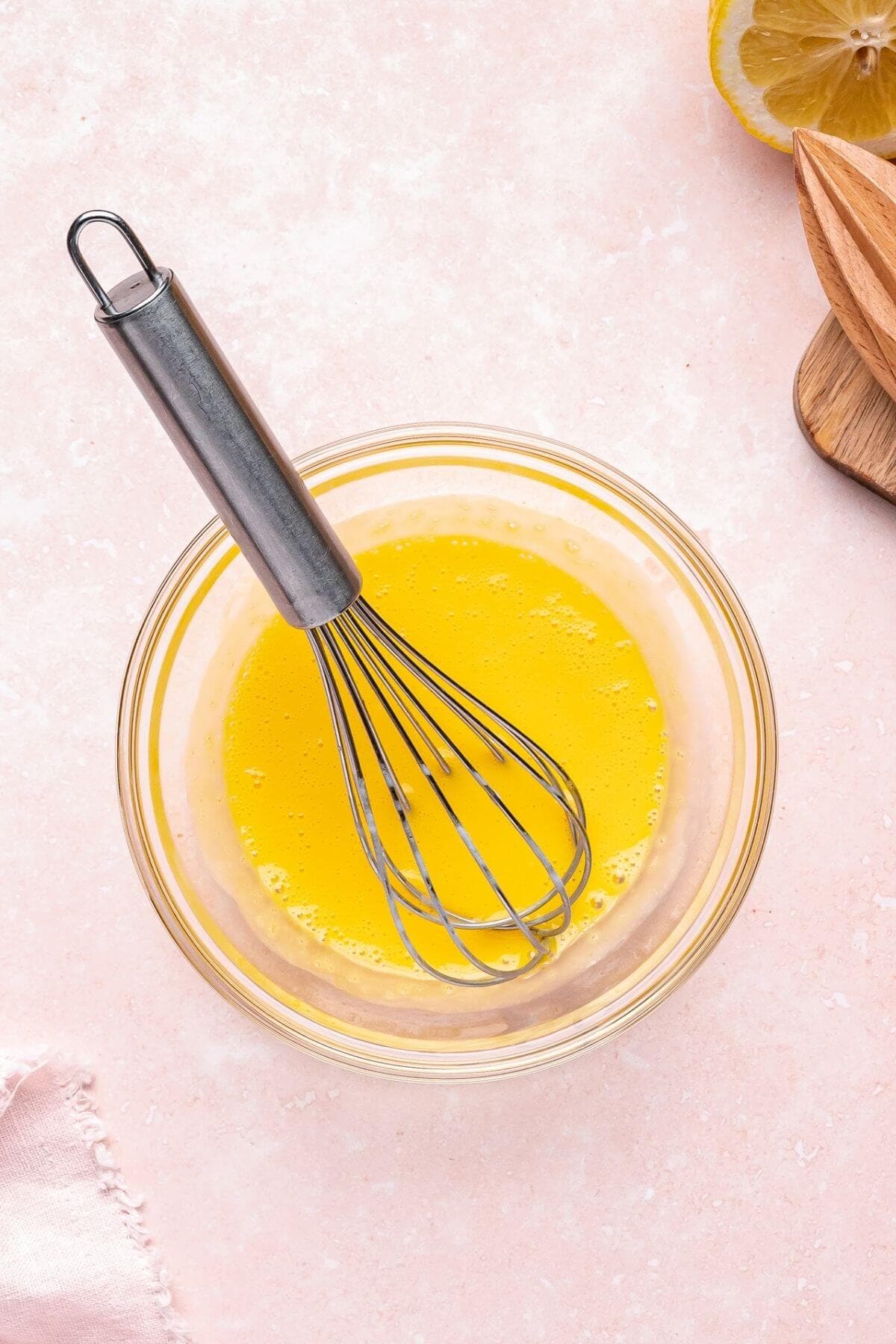 A metal whisk rests in a glass bowl containing beaten eggs on a light pink countertop, with part of a lemon and a wooden juicer visible nearby.