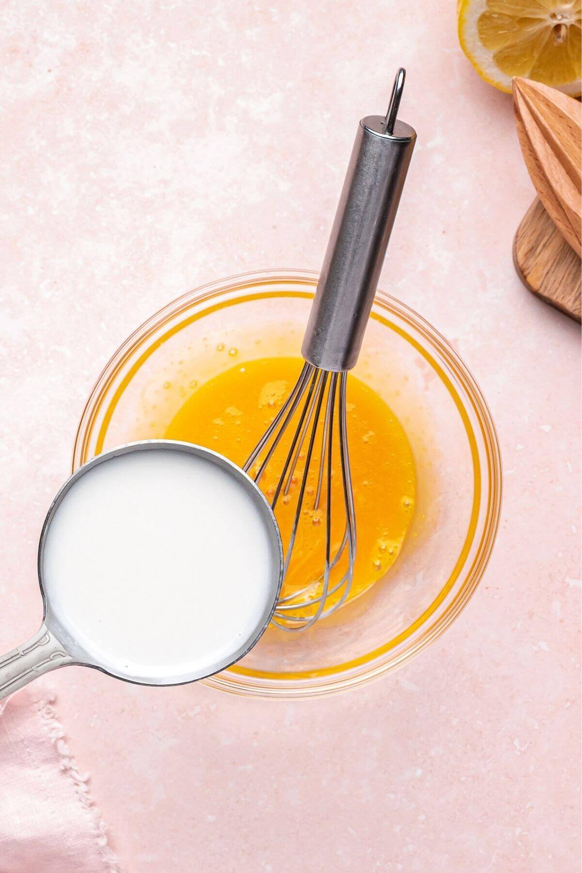 A metal whisk in a bowl of beaten eggs, with a measuring cup of milk being poured in; a lemon and juicer are partly visible on the side.