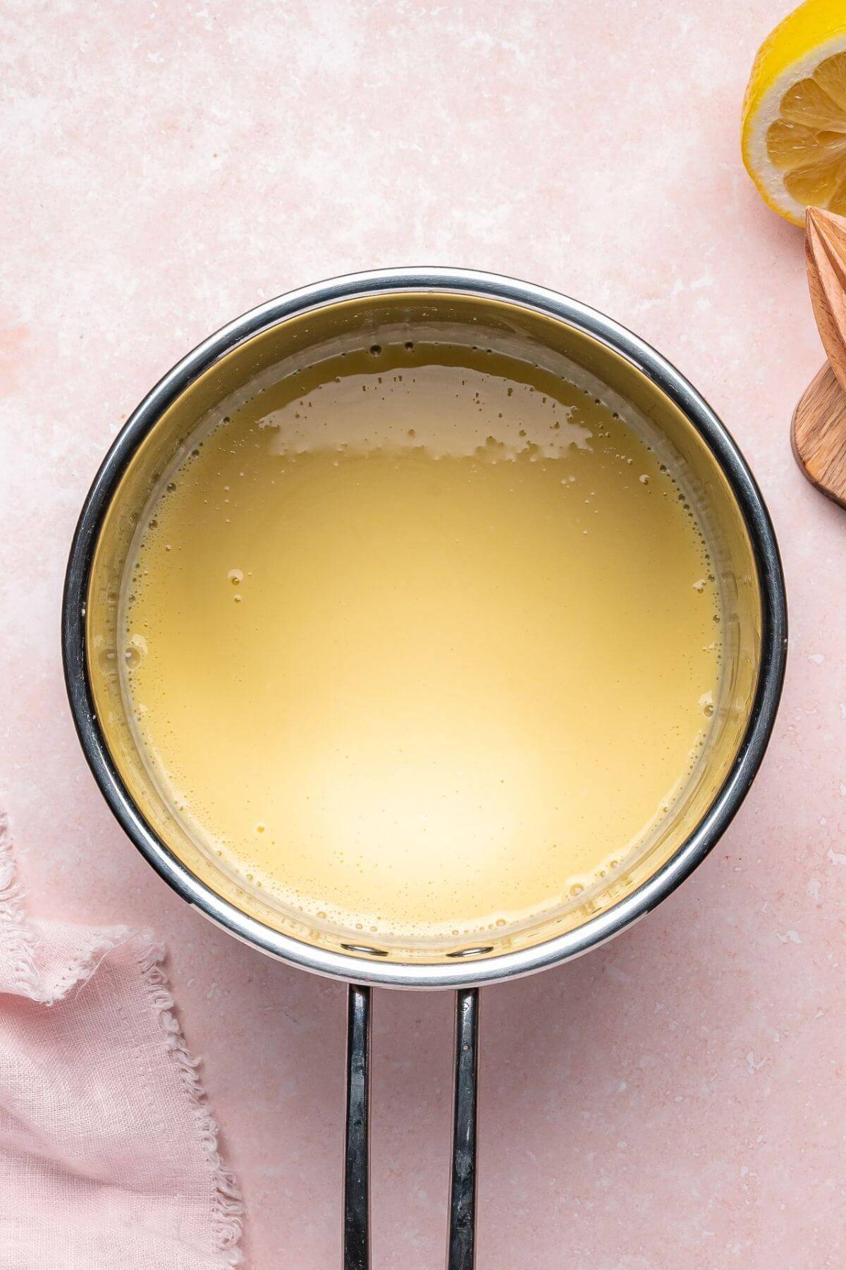 A small saucepan filled with a pale yellow liquid sits on a pink surface next to a lemon wedge, a wooden spoon, and a pink cloth.
