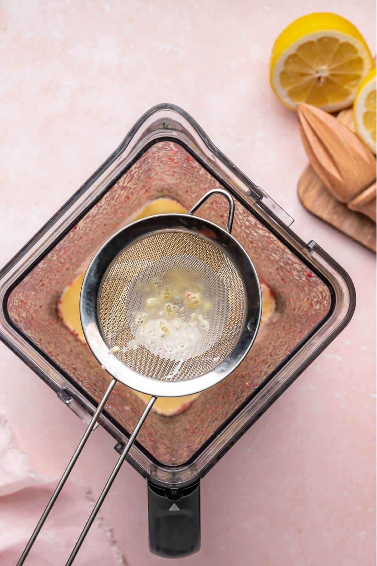 A metal strainer with lemon juice is held over a blender containing a mixture, with a halved lemon and a wooden juicer on the side.
