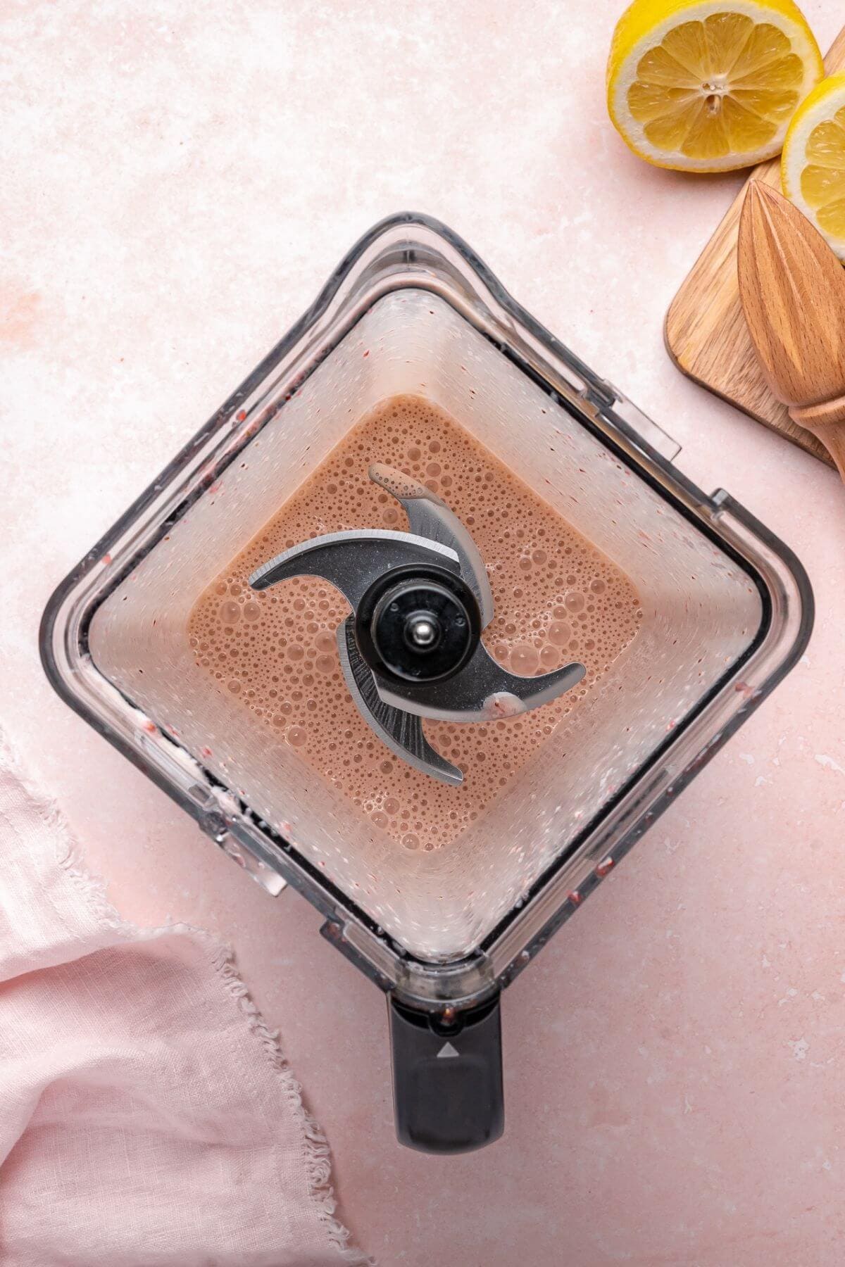 Overhead view of a blender with light brown liquid inside, surrounded by lemon halves, a wooden spoon, and a pink cloth on a light pink surface.