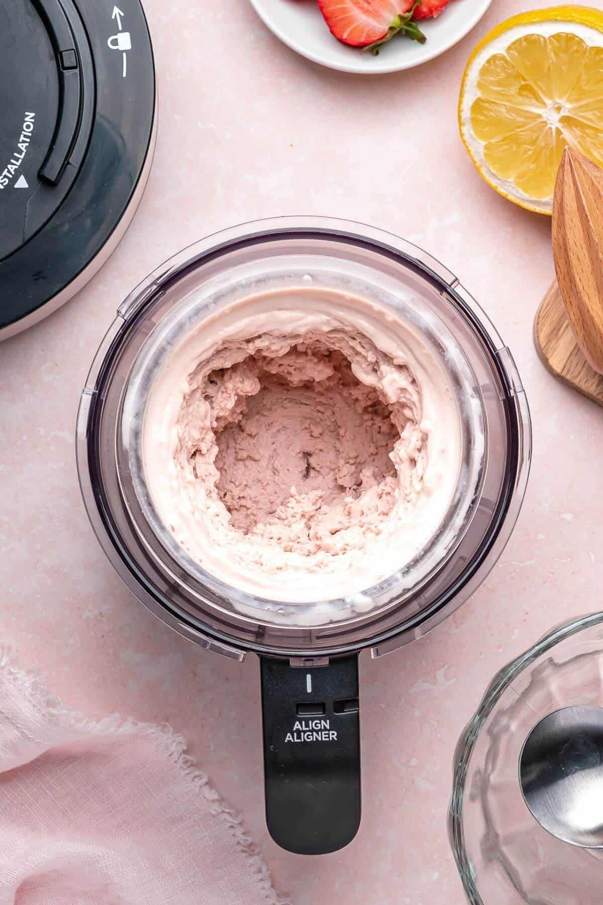 A food processor bowl contains mixed pink strawberry ice cream, surrounded by a sliced lemon, strawberries on a plate, and various kitchen items on a pink surface.