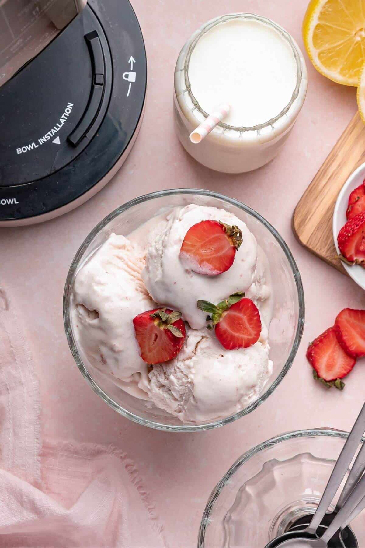 A glass bowl filled with scoops of strawberry ice cream topped with fresh strawberries, beside a glass of milk, lemon slices, sliced strawberries, and a blender base.