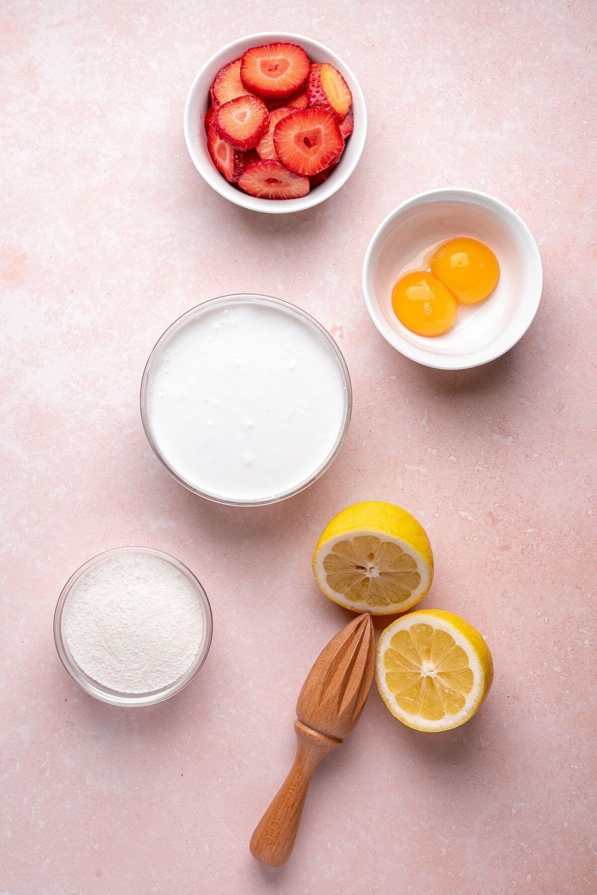 Five ingredients on a pink surface: sliced strawberries, two egg yolks in a bowl, a bowl of cream, a bowl of flour, and a halved lemon with a wooden citrus juicer.