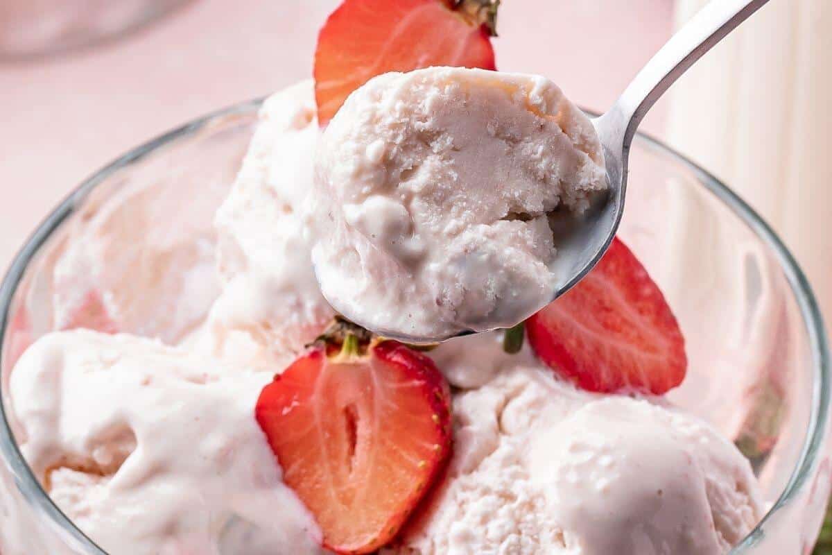 A close-up of a spoonful of strawberry ice cream in a glass bowl, garnished with fresh strawberry halves.