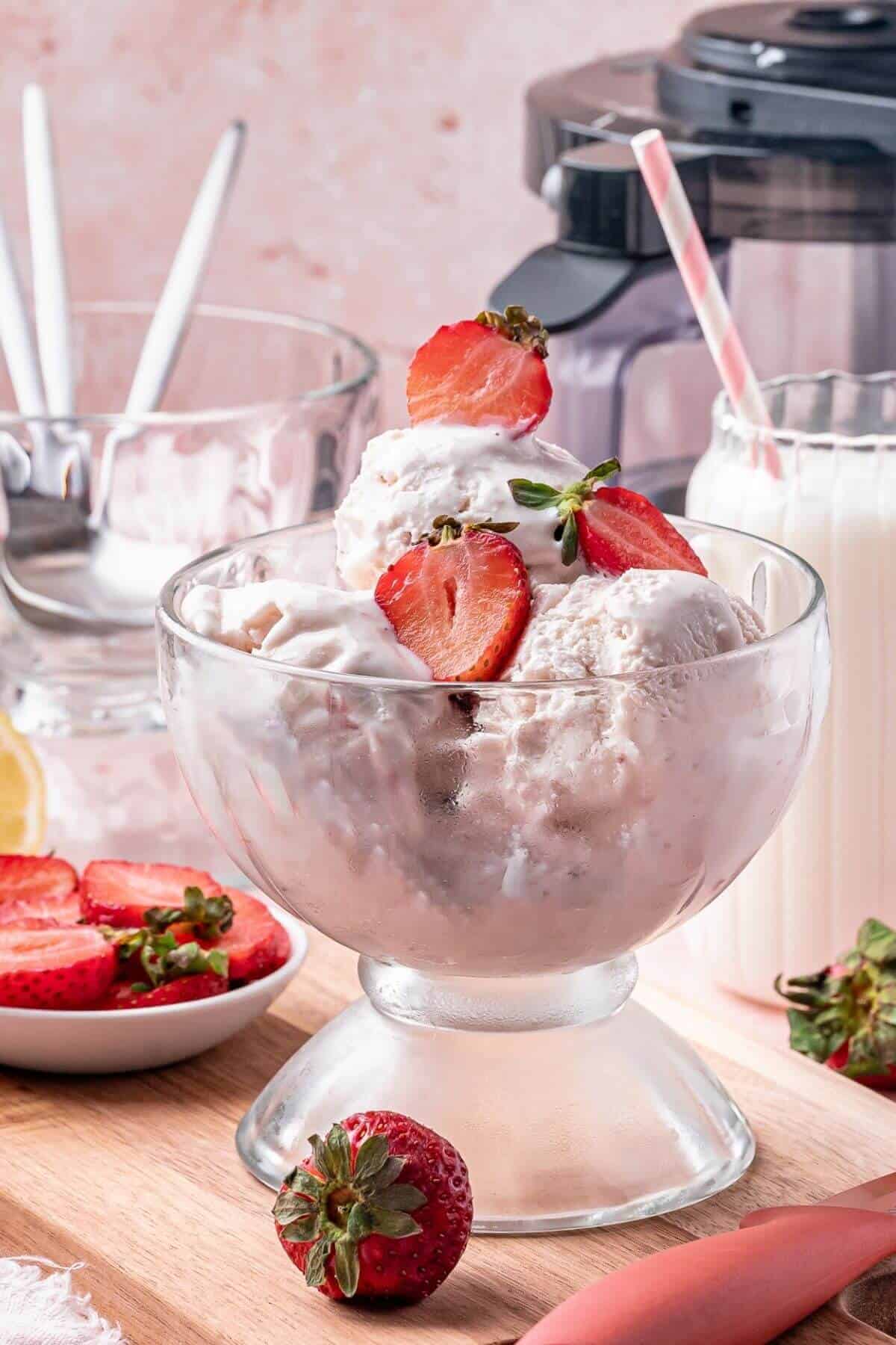 A glass bowl filled with scoops of strawberry ice cream, topped with fresh strawberry slices, sits on a wooden surface with milk, strawberries, and utensils in the background.