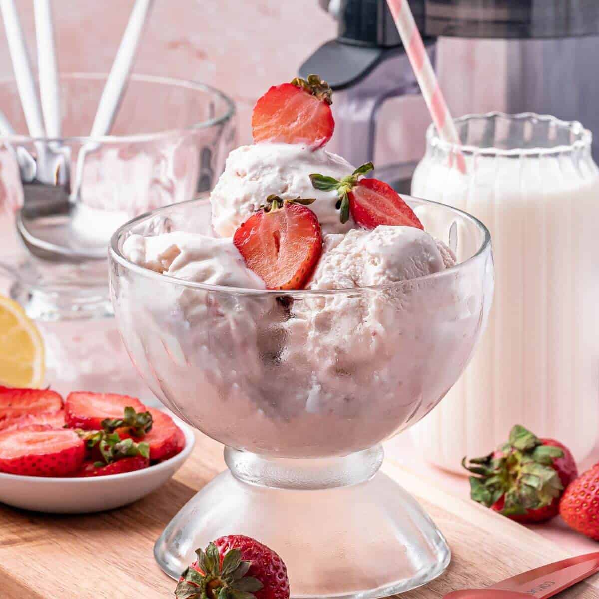 A glass dish filled with scoops of strawberry ice cream, topped with fresh strawberry slices, sits on a wooden surface with whole strawberries and a glass of milk nearby.