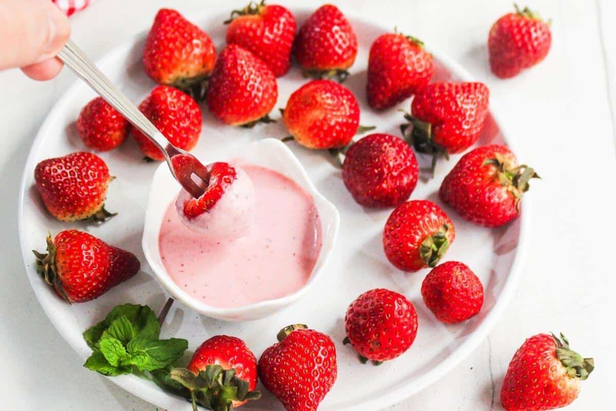 A plate of fresh strawberries surrounds a small bowl of pink dipping sauce, with a hand holding a fork dipping a strawberry into the sauce. Mint leaves are placed on the plate.