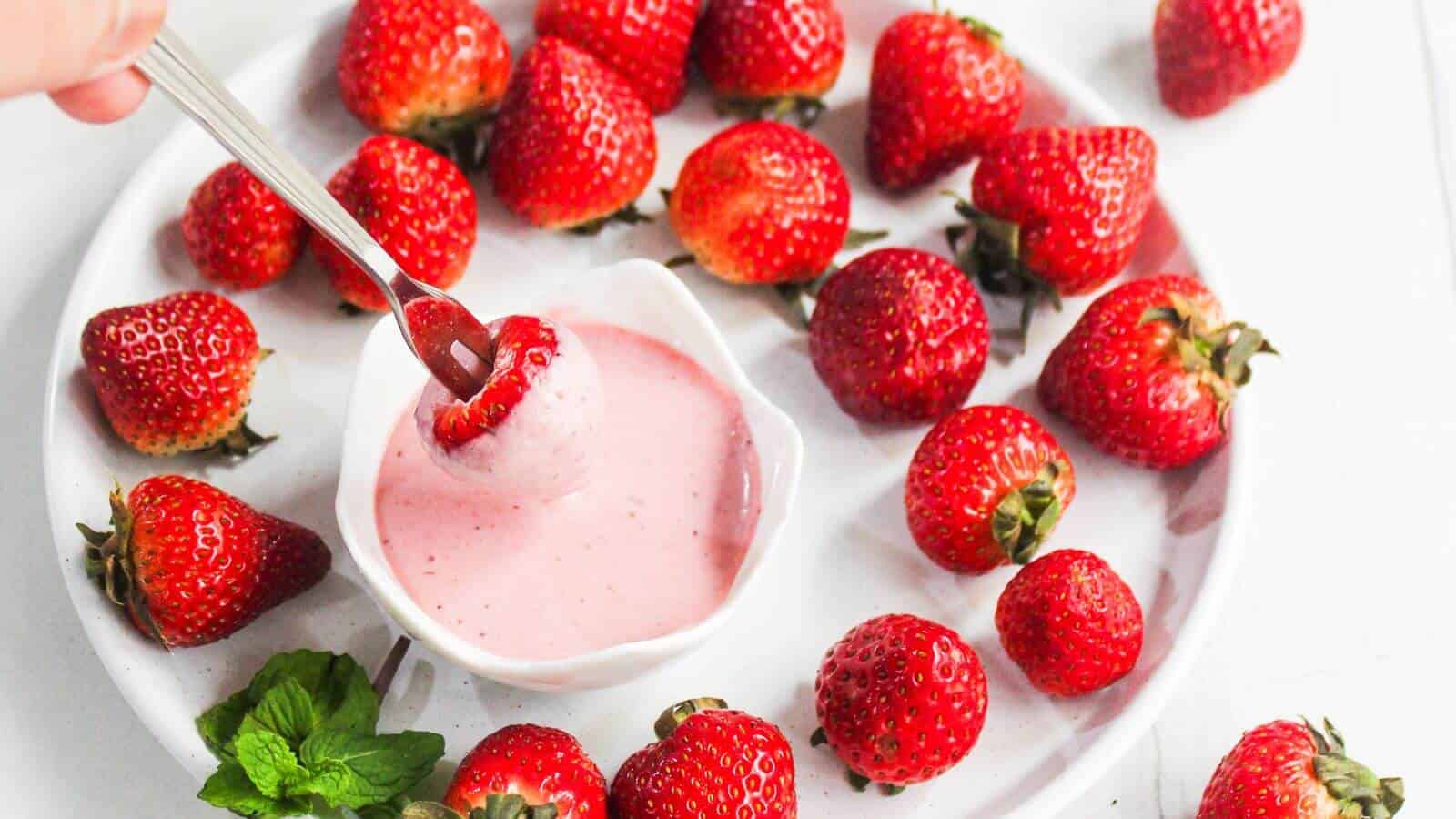 A plate of fresh strawberries surrounds a bowl of pink yogurt dip, with a hand dipping a strawberry into the yogurt.