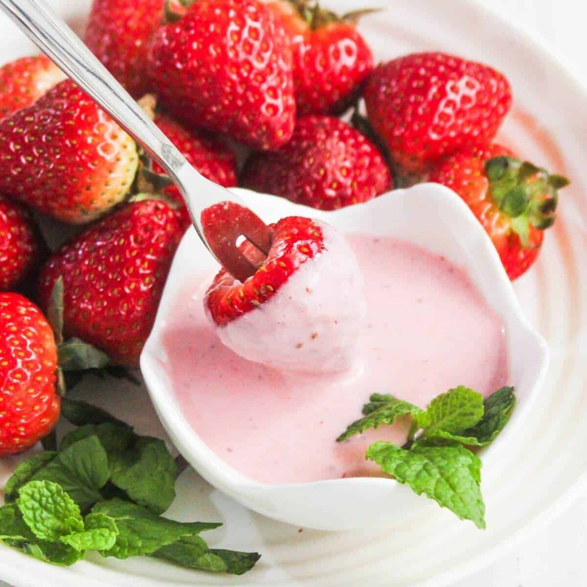 A fresh strawberry being dipped into a bowl of creamy pink yogurt, with more strawberries and mint leaves on a white plate in the background.