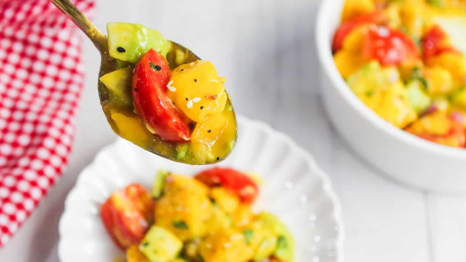 A spoonful of colorful mango avocado salad is held above a white plate, with a bowl of the same vibrant salad in the background.