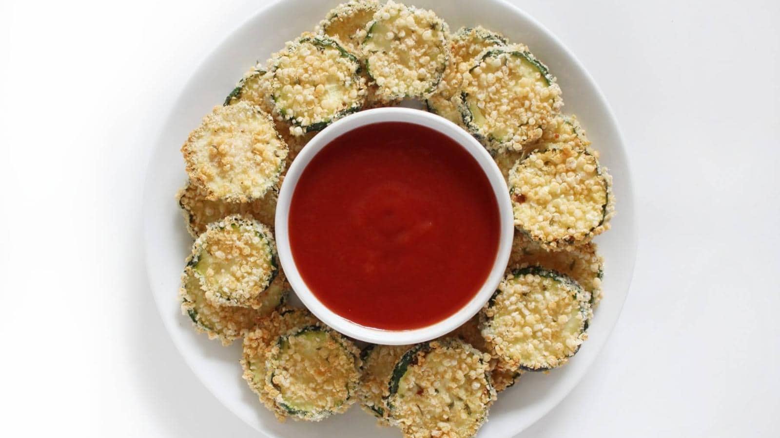 A white plate with breaded zucchini slices arranged in a circle around a bowl of red dipping sauce.
