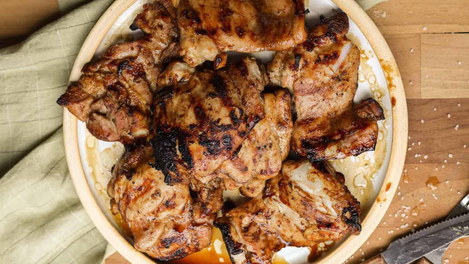 A plate of grilled chicken thighs on a round dish, placed on a wooden surface with a green cloth and scattered salt nearby.