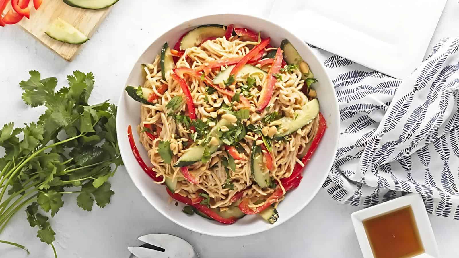 A bowl of noodle salad with sliced cucumbers, red bell peppers, and chopped peanuts, garnished with cilantro, on a white surface with a striped napkin and a small bowl of sauce nearby.