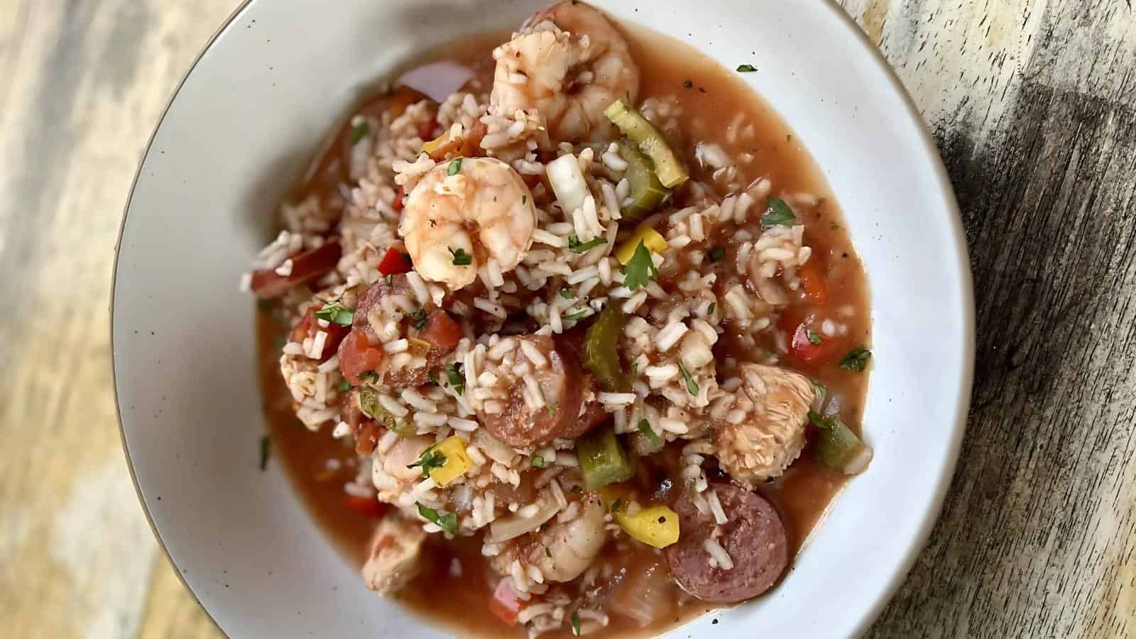 A bowl of jambalaya with shrimp, sausage, chicken, rice, and vegetables in a tomato-based sauce on a wooden surface.
