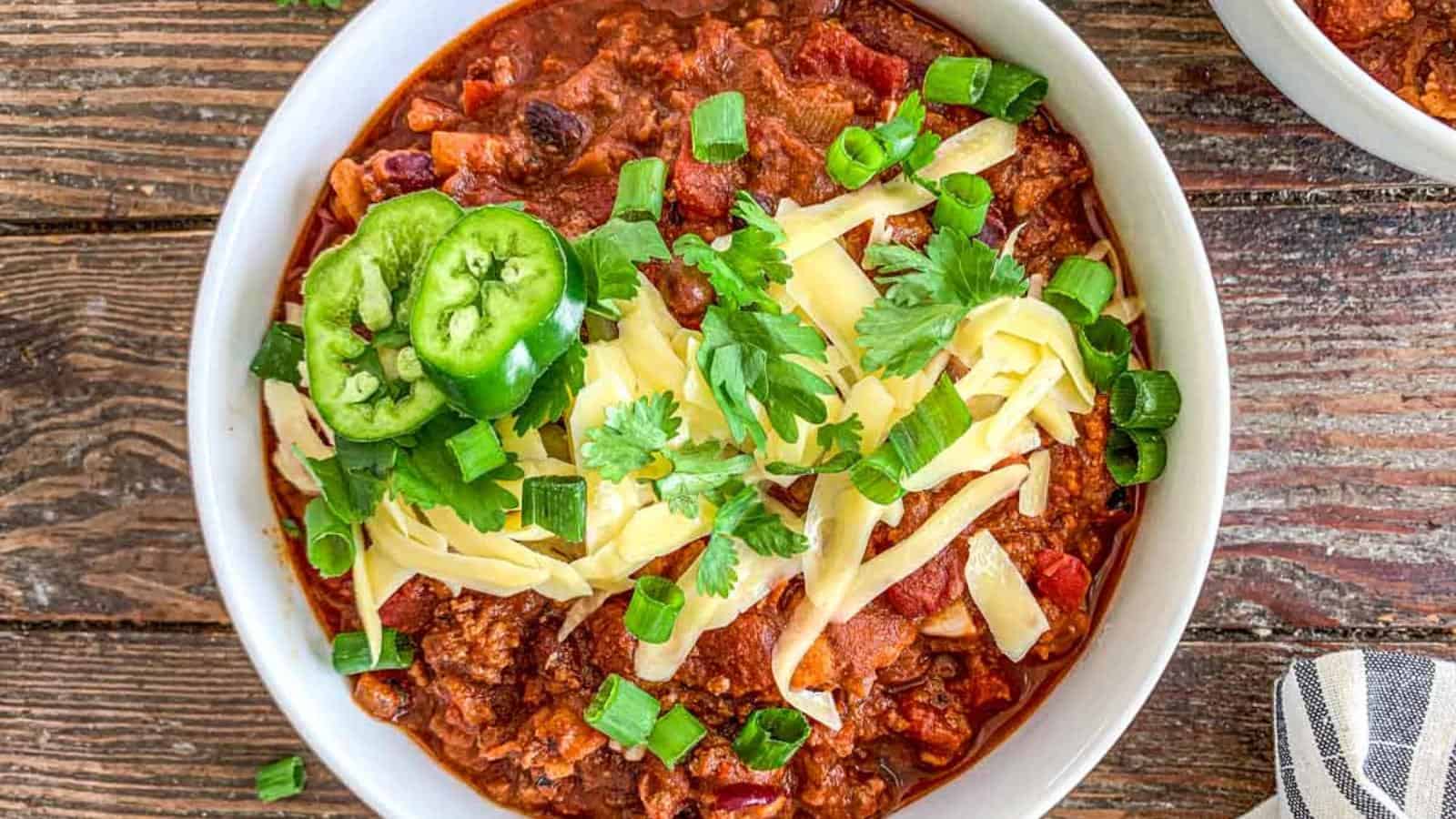 A bowl of chili topped with shredded cheese, sliced jalapenos, chopped green onions, and cilantro on a wooden table.