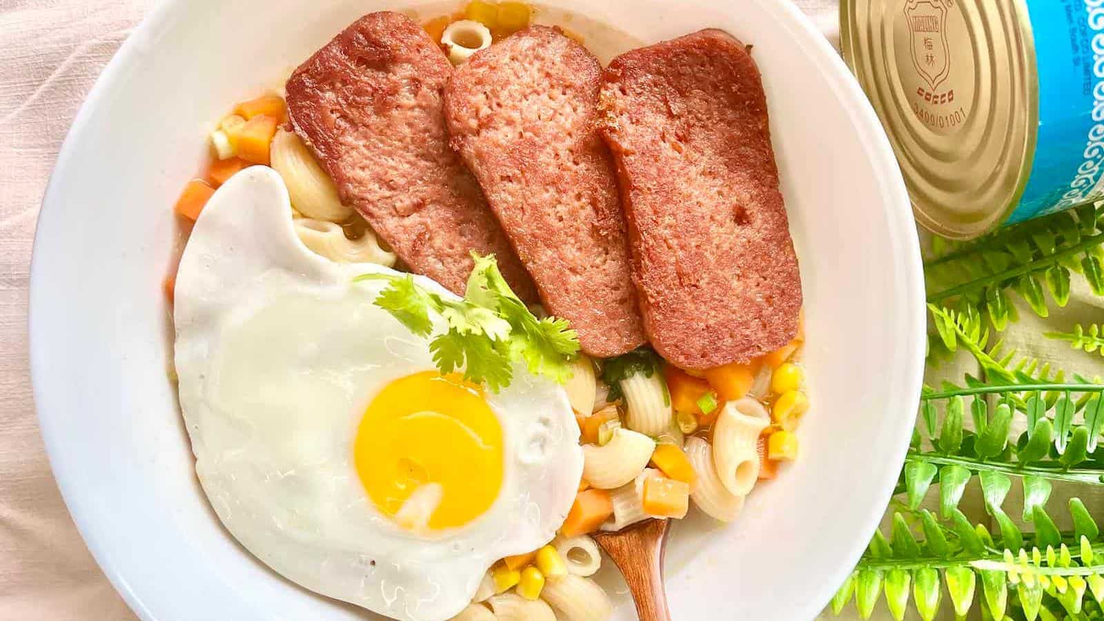 A bowl of macaroni soup topped with three slices of luncheon meat, a sunny side up egg, mixed vegetables, and garnished with cilantro. A can and green plant are beside the bowl.